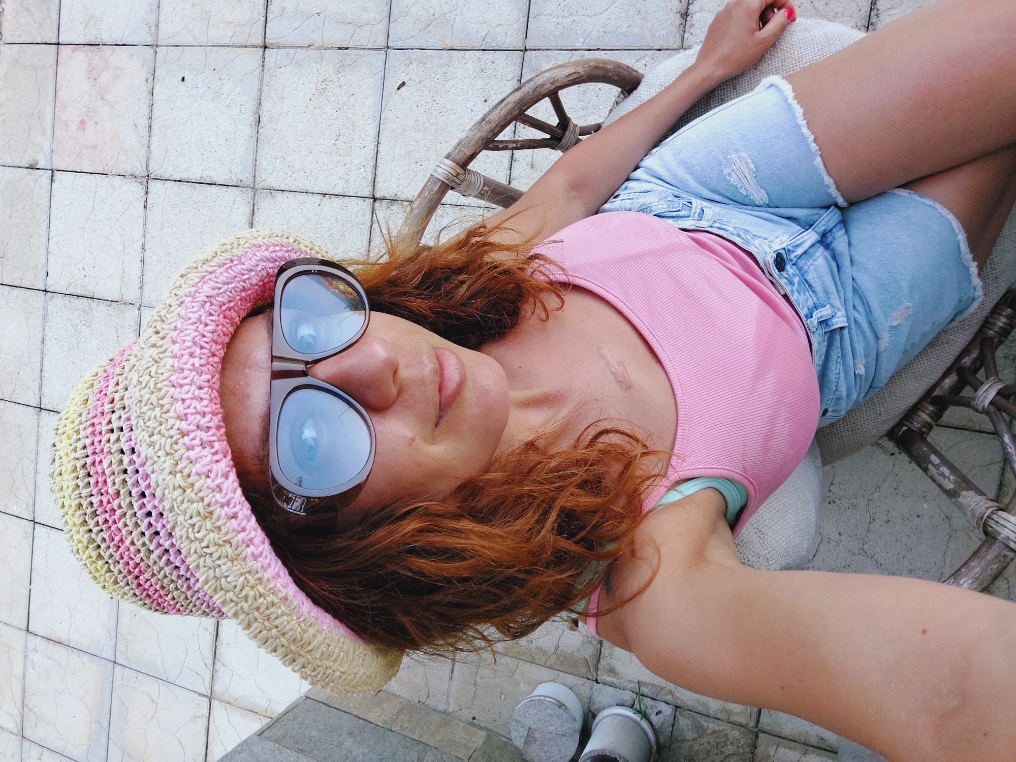 A woman is taking a selfie while sitting on a chair outdoors, wearing a pink tank top, denim shorts, a straw hat, and sunglasses. The woman has red hair and is wearing light blue denim shorts with distressed details. The background shows tiled flooring and what appears to be a stone wall or structure. The scene is likely in an outdoor seating area.