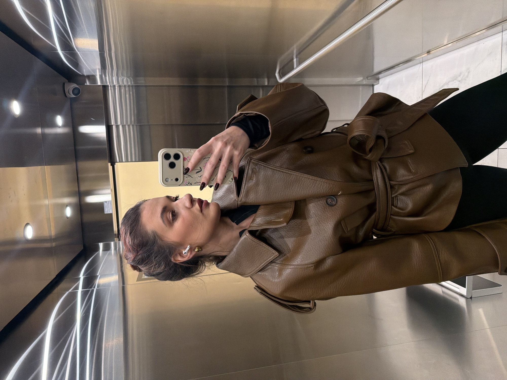 A female individual is depicted taking a mirror selfie within an elevator interior in Belgrade, Serbia. The subject, centrally positioned, wears a brown, belted, double-breasted trench-style jacket over a black long-sleeved top and black leggings. Her brown hair, with subtle reddish undertones, is styled in an updo. A gold hoop earring and a white wireless earbud are visible in her left ear. Her fingernails are painted a dark red. She holds a light-colored smartphone, featuring a multi-lens camera system and a floral-patterned case, in her right hand, directing it towards her reflection. Her gaze is directed towards the phone screen. The elevator features polished metallic walls, likely stainless steel, reflecting ambient light and the subject. Recessed circular LED lights are mounted on the ceiling, where a white dome-shaped surveillance camera is also visible in the upper right. A silver horizontal handrail runs along the right wall. The floor is tiled with white, possibly marble, squares. A small, rectangular metallic fixture is mounted on the lower left wall.