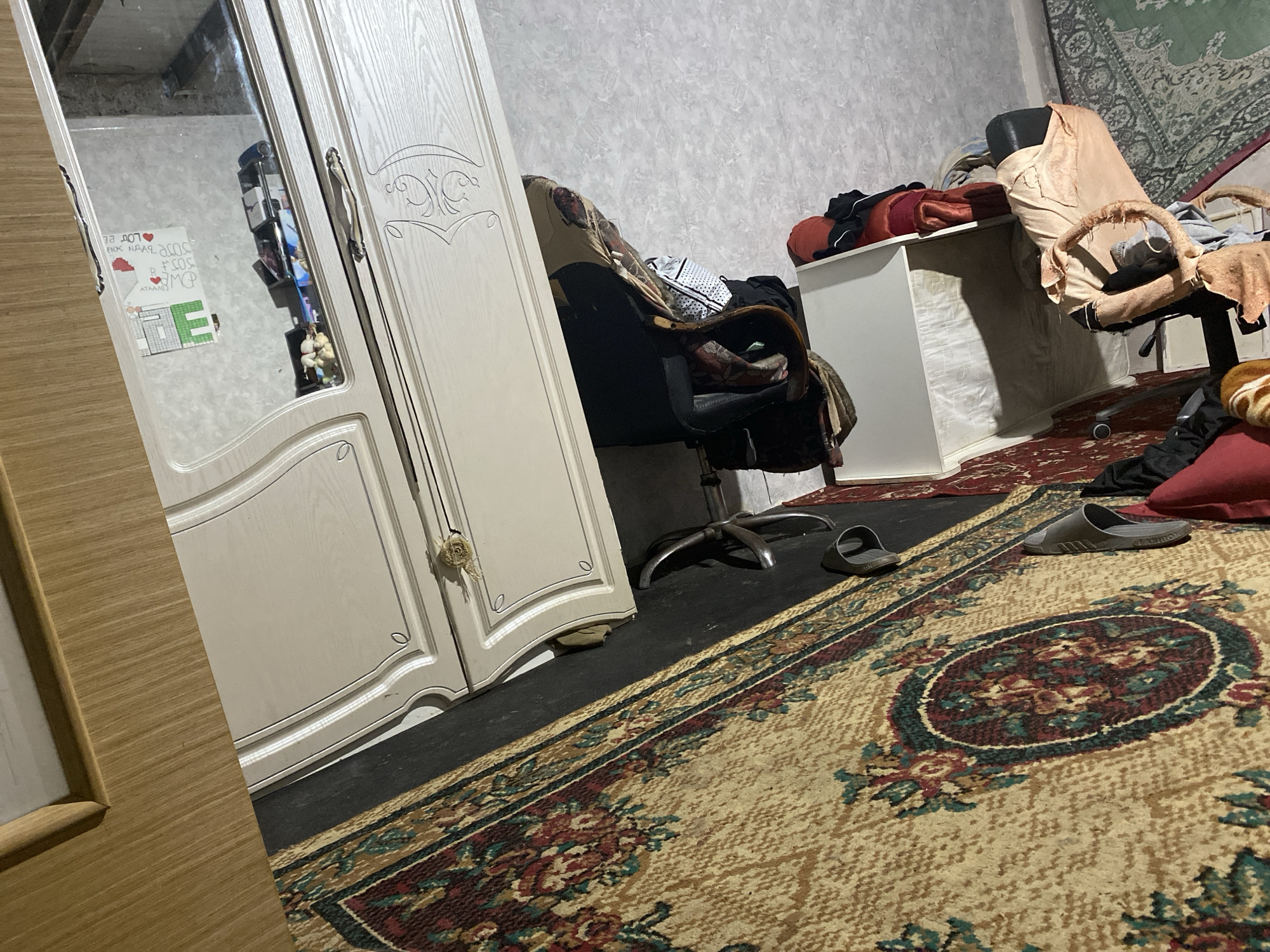 The image depicts an interior scene, likely a room in a residence located in Shakhtinsk, Kazakhstan. The room appears cluttered, with various items strewn about. In the foreground, a large, patterned rug covers a portion of the floor. A pair of gray slide sandals are visible on the rug. To the left, a tall, light-colored wardrobe with mirrored doors is present. The wardrobe's surface is decorated with carved patterns. On the floor in front of the wardrobe, a dark mat is visible. Further back, an office chair is positioned next to a white desk. The chair is draped with fabric, and a pile of clothing is on the desk. A red pillow or cushion is also on the desk. In the background, a green rug with an intricate pattern hangs on the wall. The walls themselves have a textured wallpaper. The lighting in the room appears dim, casting shadows.