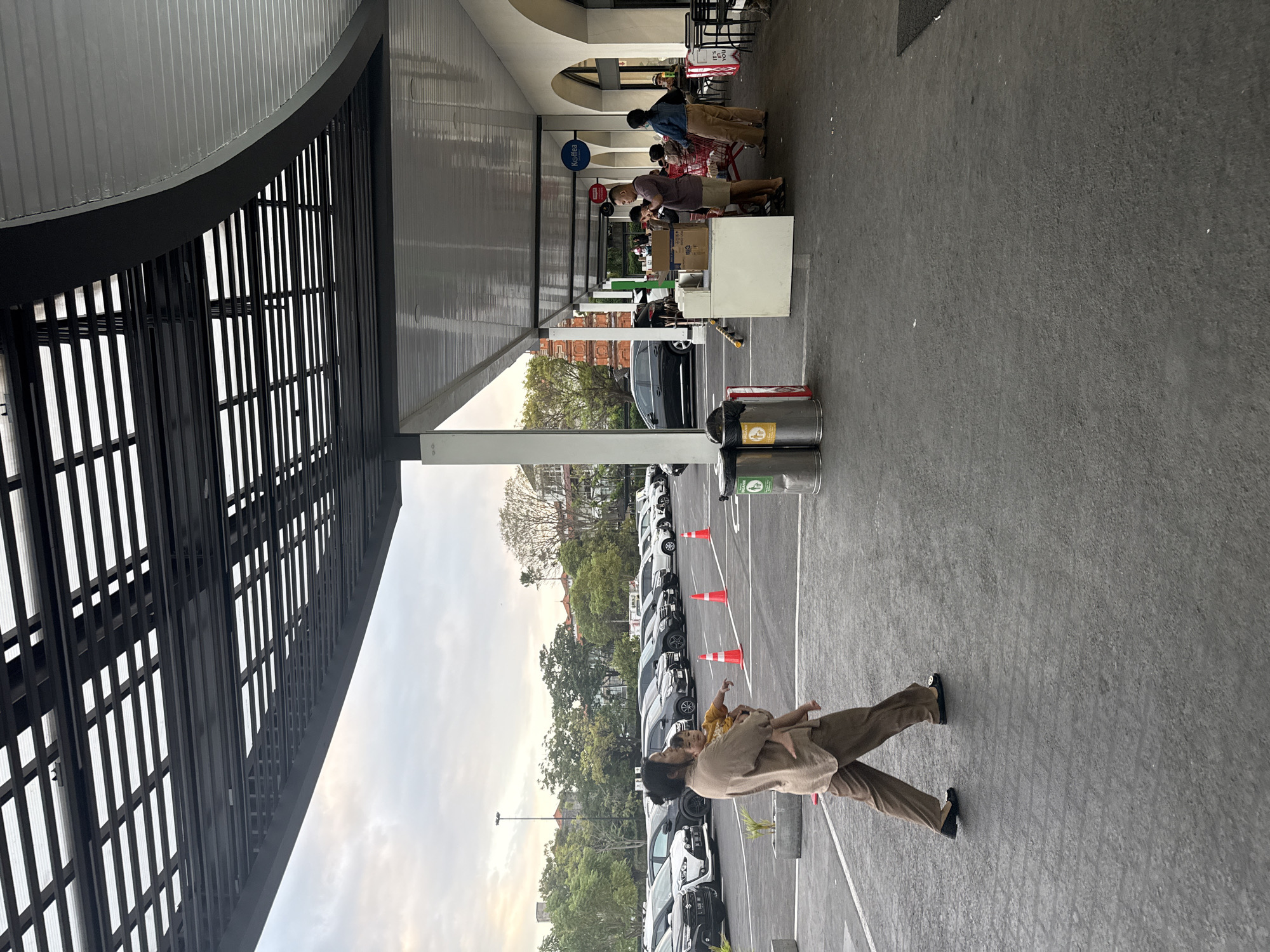 A woman wearing a loose-fitting brown outfit and black flats walks across a paved lot, holding a baby in her arms. The baby is wearing a yellow shirt. They are walking in front of a row of parked cars. The lot appears to be a parking area in front of a building with a covered walkway. The walkway has a modern, dark-colored slatted roof structure. Several people are gathered under the walkway, some standing around a counter with boxes, and others seated at tables outside a cafe labeled "Koffea". The sky is overcast. The scene is likely a commercial area in Denpasar, Indonesia, given the presence of parked cars that are common in the city and the architectural style which could be found in modern establishments in the region.