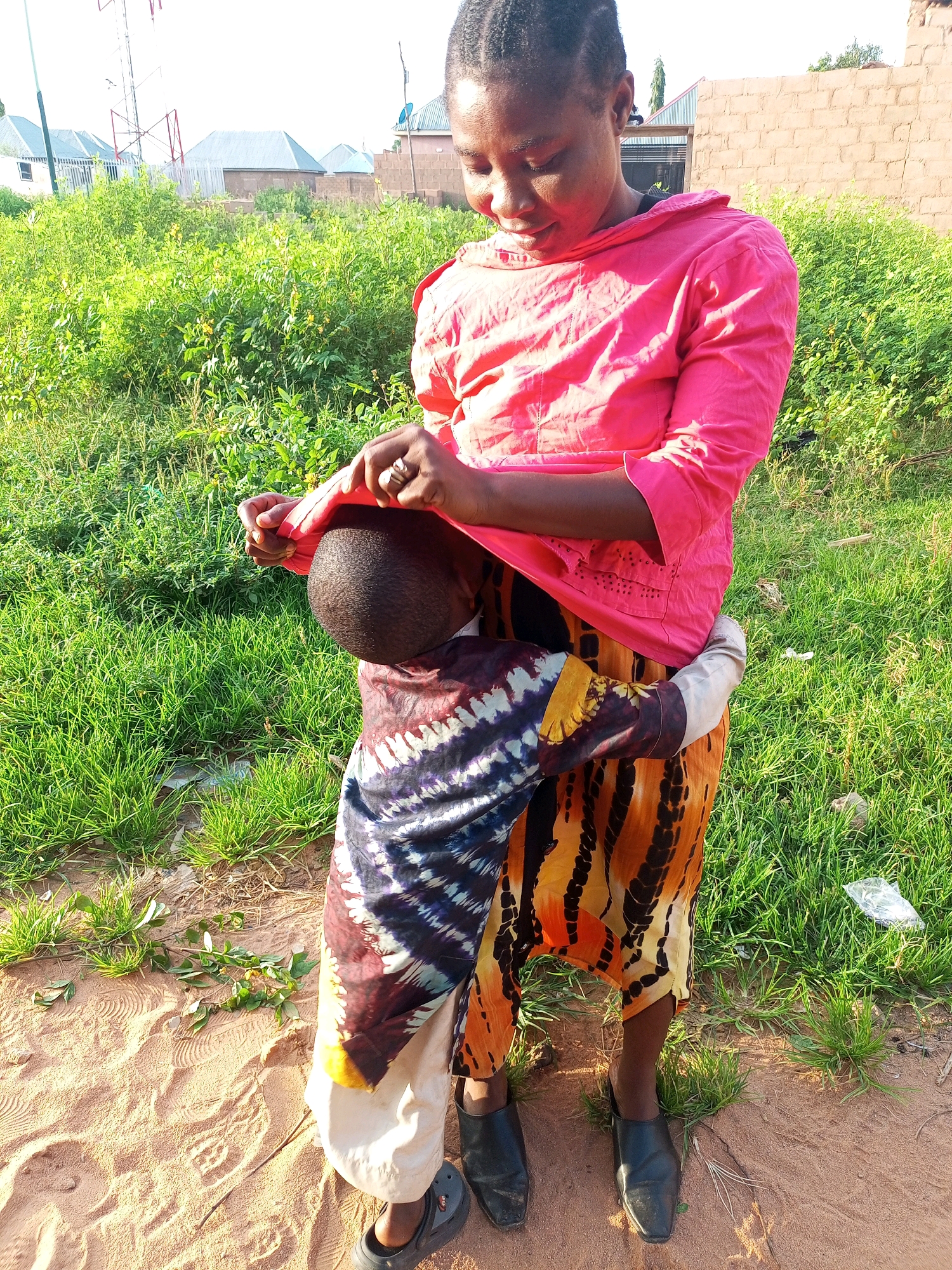 A young woman in a bright pink shirt and a patterned skirt is holding a young child. The child is facing away from the camera, with their head nestled against the woman's chest. The woman is looking down at the child with a gentle smile, her finger pointing to the child's head. Both are wearing traditional clothing. The scene is outdoors, with lush green grass and sandy ground. In the background, there are some buildings and a tall telecommunications tower. The lighting suggests it is daytime, likely late afternoon given the warm glow. The overall emotion conveyed is one of warmth and affection.
