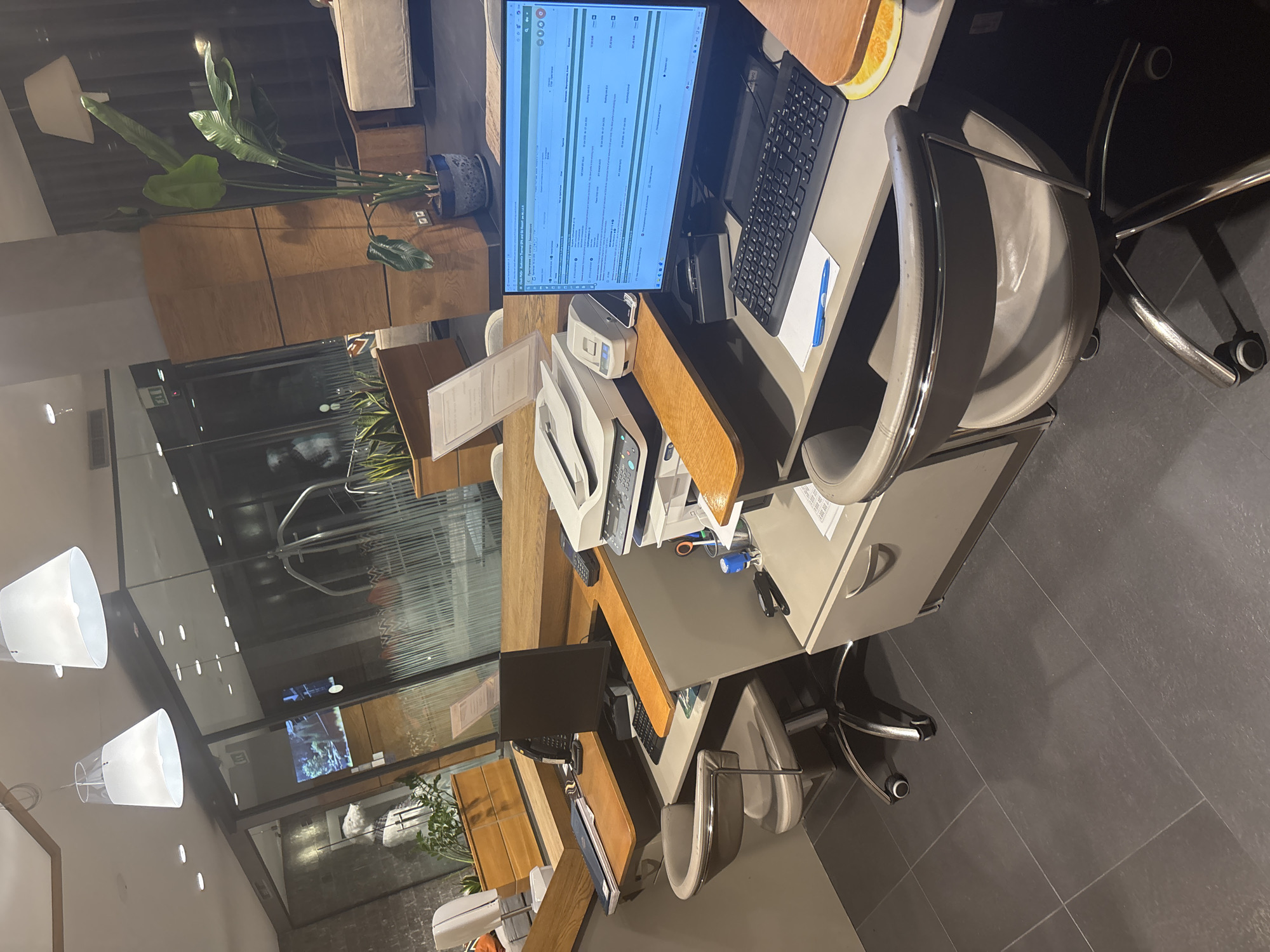 The image captures an indoor office or reception area, likely within a commercial establishment in Dobrinishte, Bulgaria. The space is unoccupied, presenting an orderly and functional atmosphere typical of a front office or administrative workstation.
The central feature is a desk area comprising a wooden surface and grey filing cabinets. A computer monitor displays what appears to be a booking or inventory management system, showing a tabular interface with columns labeled "Item," "Event," "Date," "Status," and "Price." Visible entries include "Test Reservation," dates like "13.08.2021," "02.04.2023," and "28.02.2023," alongside prices, including "115.00 EUR." A search bar reads "Search reservations / guests..." A keyboard, mouse, and a blue pen on a mousepad are positioned in front of the monitor. Adjacent to the desk, a white multi-function printer/scanner is visible, and on a lower shelf, various office supplies such as remote controls, scissors, and a pen holder are organized. A grey upholstered desk chair with a chrome frame is placed at the workstation, and a distinct orange-colored circular coaster or decorative item rests on the desk.
Adding a natural element to the interior are large potted plants with broad green leaves, possibly a Bird of Paradise, situated behind the main desk. The background features wooden wall panels or cabinetry, and a reflective glass partition offers a glimpse into another interior space. Two modern white conical pendant lights illuminate the area from above. The floor is tiled with large, dark grey squares. A partial view of another similar workstation with a monitor and chair can be seen in the lower foreground. The overall impression is one of a well-maintained and ready-to-use professional environment.