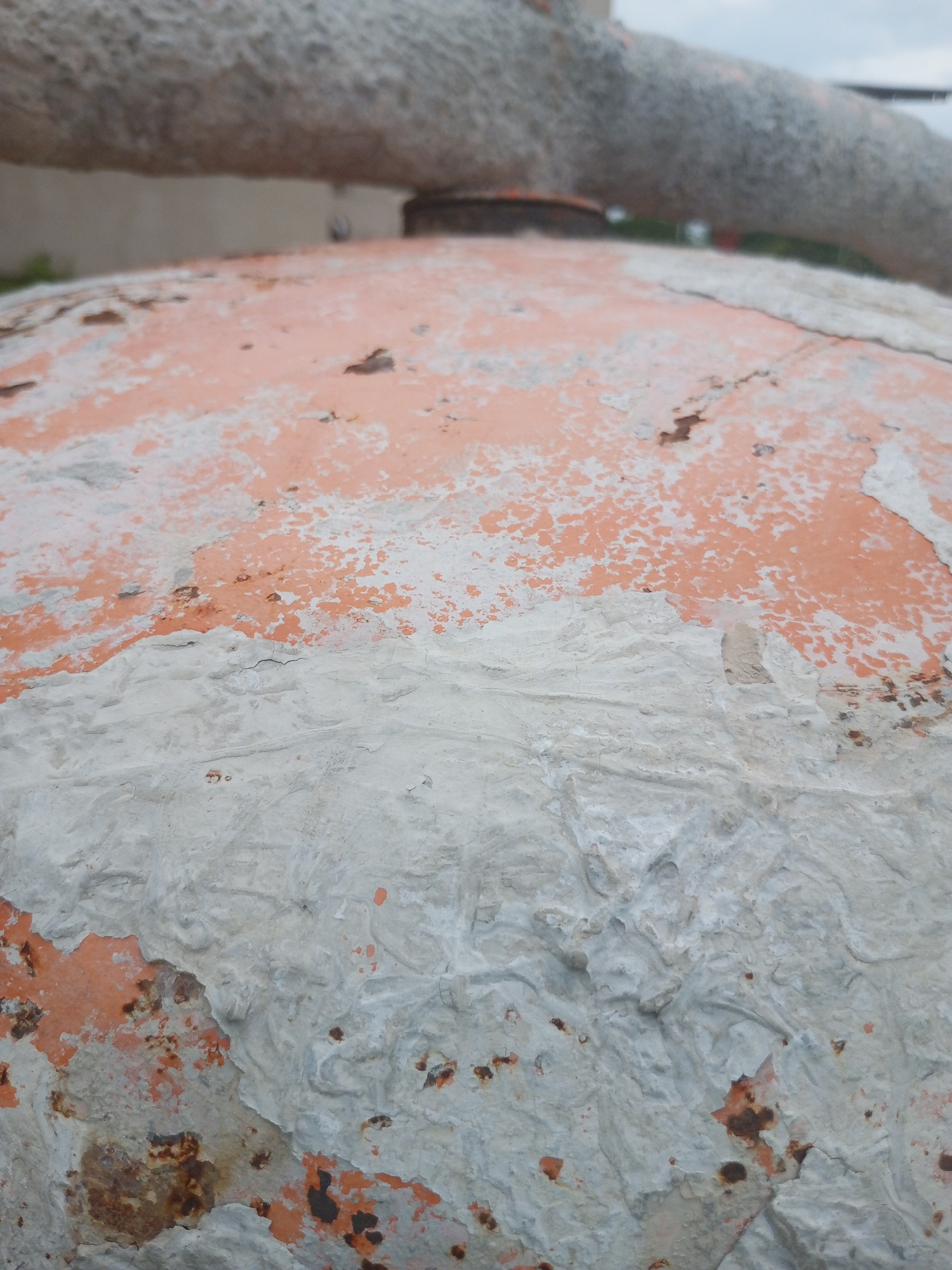 The media file is a close-up, low-angle shot of a weathered, curved surface, likely a tank or similar industrial object, with peeling orange and white paint. The surface has a textured, rough appearance due to the deteriorated paint and possibly rust. There are hints of a grey, textured object in the upper portion of the frame, which could be part of the same structure or a connected element. The background is out of focus, showing a blurred suggestion of an outdoor setting with greenery and a muted sky, suggesting it is daytime. The overall impression is one of age and disuse. No people, text, or specific event are discernible. The context provided places this scene in Jalingo, Nigeria.