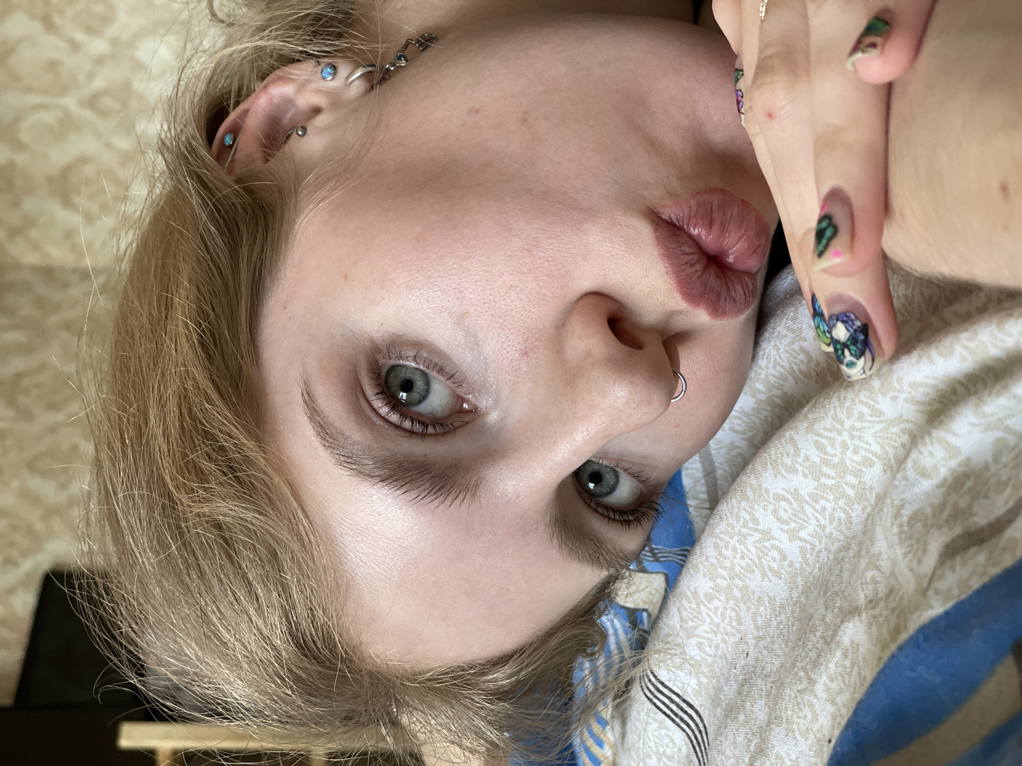 A close-up, rotated, indoor shot depicts a young woman with fair skin, blonde hair, and light-colored eyes. She is lying on a bed with a patterned duvet. A silver nose ring adorns her left nostril, and multiple piercings are visible on her left ear. She holds her hand up, revealing painted fingernails with butterfly designs. The location or presence of Izhevsk, Russia, is not discernable from this image.