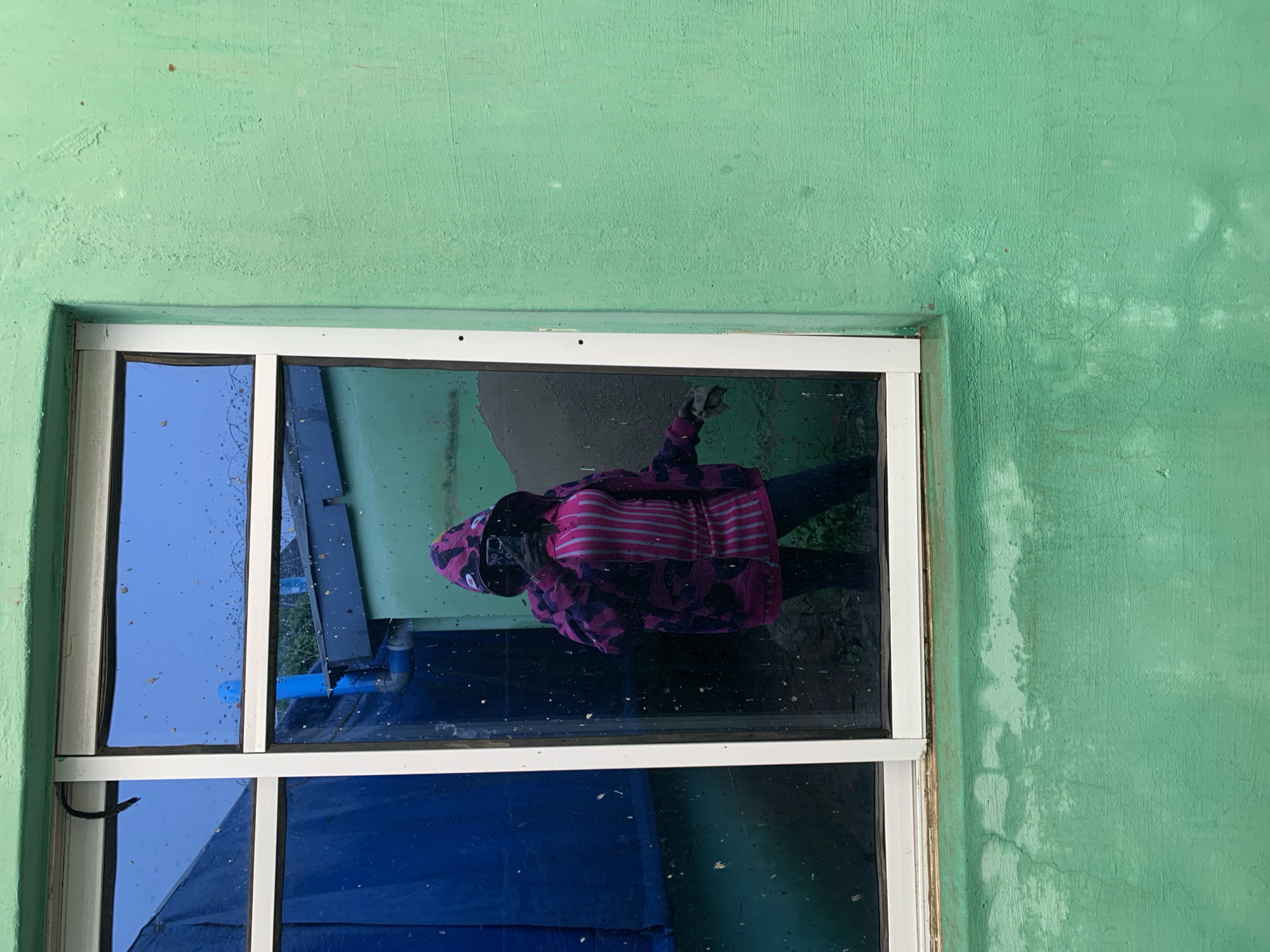 The image shows a close-up view of a window on a green, textured wall. The window is divided into four panes by white frames. The glass in the window is reflective, showing a distorted reflection of a person wearing a hooded sweatshirt with a pink and purple camouflage pattern and pink stripes. The person appears to be holding a phone, likely taking a selfie. The reflection also shows some blue structures and greenery outside. The wall outside the window has some water stains or discoloration. The overall scene is an outdoor setting, possibly residential or a simple building. There is no visible text in the image. The lighting suggests it could be daytime.