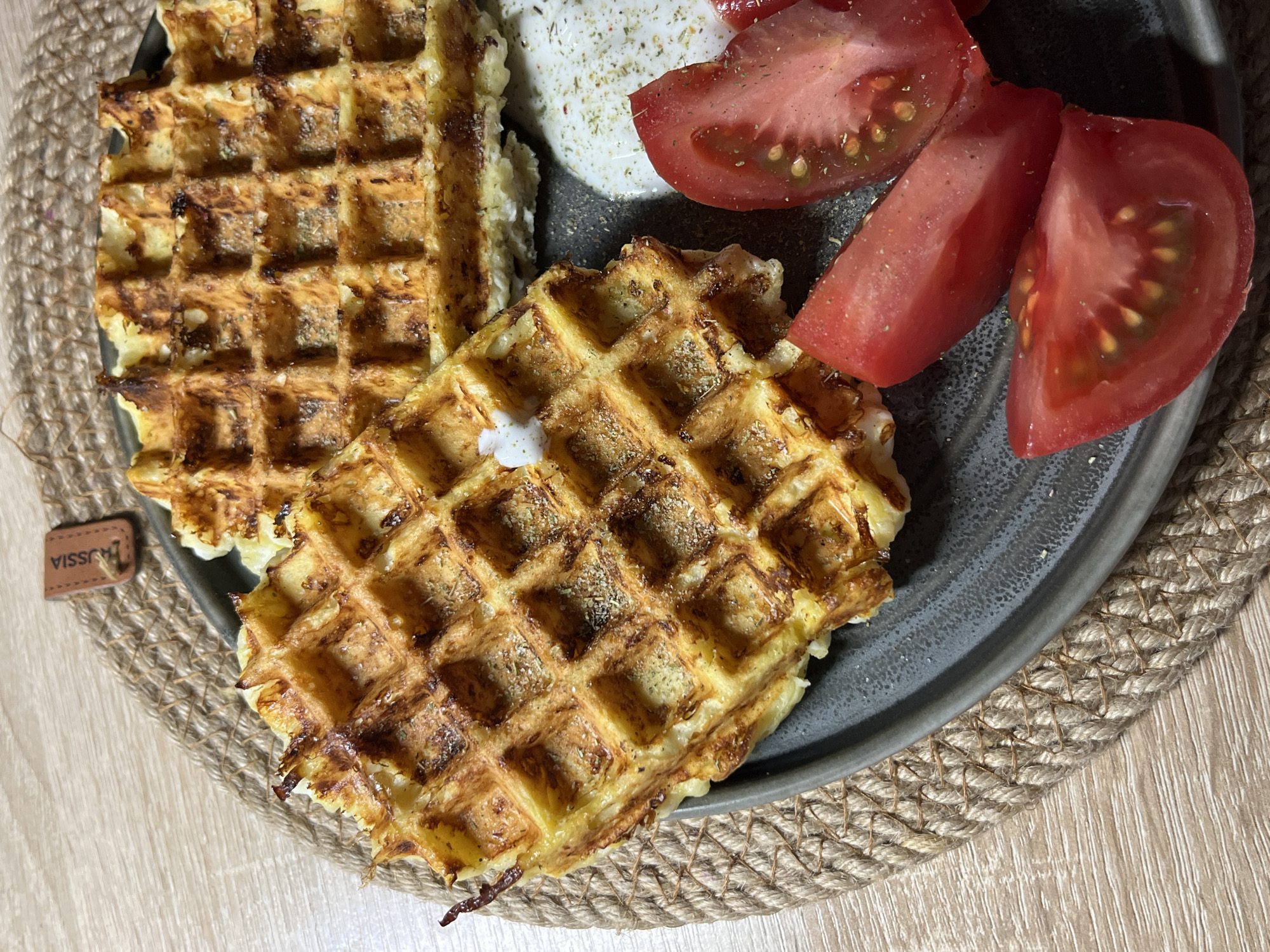 The image presents a close-up, overhead view of a savory meal laid out on a table, likely for breakfast or brunch. The main subjects are two golden-brown waffles, one partially overlapping the other, both exhibiting a crisp texture and appearing to be seasoned with dried herbs or spices. Accompanying the waffles on a dark gray, matte-finish ceramic plate are several wedges of vibrant red tomatoes and a dollop of white, creamy substance, possibly a cheese or yogurt sauce, both also garnished with dried green herbs. A small piece of the white cream is visible on one of the waffles.
The plate is positioned on a round woven placemat, made of natural, light brown cord or rope, which contrasts with the smooth, light-colored wooden surface beneath. The setting suggests an indoor environment, illuminated by bright, diffused light, indicating daytime.
A notable detail is a small, brown, leather-like tag or label situated on the placemat to the left. The word "JUSSIA" is clearly visible in white capital letters on this tag. While no specific activities beyond the presentation of the meal are depicted, the arrangement suggests readiness for consumption. There are no people visible, nor are there explicit visual cues in the scene itself to confirm the provided location context of Sochi, Russia, other than the potential relevance of the "JUSSIA" tag to that region.