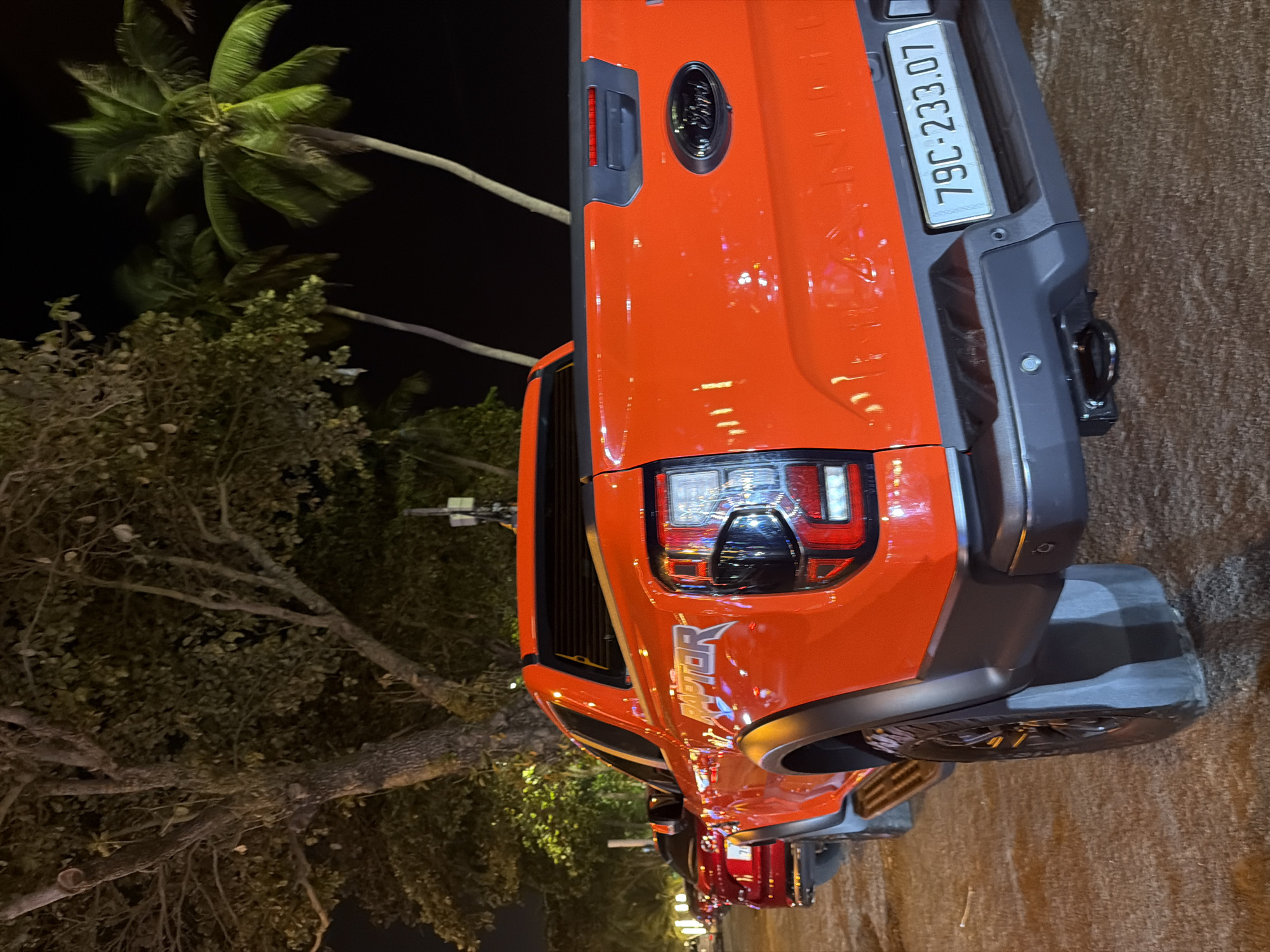 A bright orange Ford Ranger Raptor pickup truck is parked on a textured surface, possibly gravel or coarse sand, at night in Nha Trang, Vietnam. The truck is viewed from the rear, three-quarters to the left. The license plate is clearly visible and reads "79C-233.07". The "RAPTOR" text is emblazoned on the side of the truck bed in white lettering with a silver outline. A Ford logo is present on the tailgate. The truck has prominent black fender flares and a black rear bumper.
In the background, several palm trees with lush green fronds are visible against a dark night sky. To the left of the Ford Raptor, the rear of a red car, possibly another vehicle, is partially visible. A large tree with visible branches and foliage stands to the left of the vehicles, casting shadows and illuminated by ambient light, suggesting it's located near a street or public area. The overall lighting suggests an urban or semi-urban nighttime environment, typical of coastal areas like Nha Trang. There are no people or discernable actions depicted in the image.