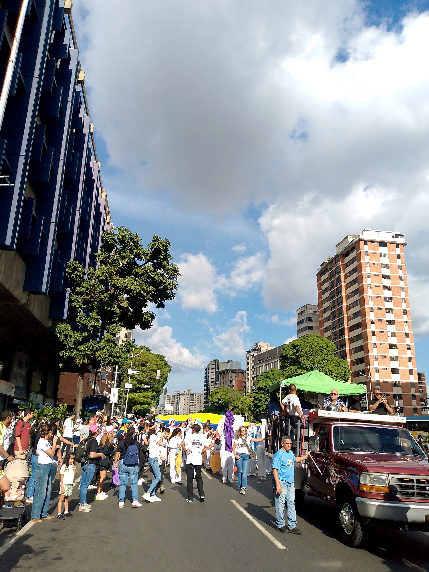 This is a wide-angle, eye-level, outdoor photograph taken on a street in Caracas, Venezuela, on a cloudy day. The scene captures a street procession or demonstration.
On the left side of the frame, a modern, dark blue building with a repetitive facade of recessed windows dominates the background. A large, leafy green tree is positioned in front of this building.
The street is filled with a crowd of people, many of whom appear to be participants in the procession. They are dressed in casual clothing, and some are holding flags or banners. The crowd is diverse, with individuals of various ages and ethnicities.
In the center of the frame, a red pickup truck is driving slowly. It has a green tent erected in its bed, providing shade for some of the individuals standing or sitting on the truck. Several people are visible on top of the truck, including one man who appears to be taking a photograph with a mobile device.
Further down the street, more buildings can be seen, including a tall, red-and-white checkered brick building. A large yellow banner is visible stretched across the street in the distance, indicating the route of the procession.
The sky is partly cloudy with patches of blue sky visible. The overall impression is of a vibrant, active urban scene during a public gathering.
