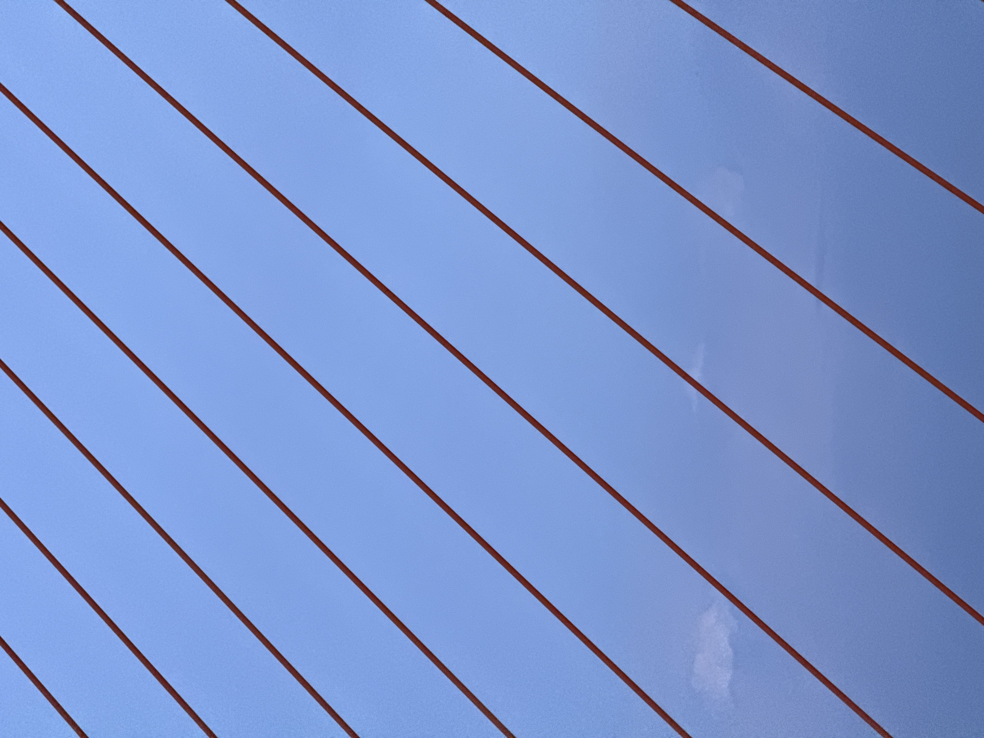 This media file depicts a close-up, abstract view of a suspension bridge's cables against a clear, light blue sky. The cables are dark red and run diagonally across the frame from the bottom left to the top right, creating a repeating parallel pattern. There are no people or specific objects visible, only the repeating geometric lines of the cables and the expansive sky. The lighting suggests it is daytime, and the clear sky implies fair weather. The context of Đà Nẵng, Vietnam, specifically points to the iconic Đà Nẵng Bridge, known for its distinctive red cables and modern design. The overall impression is one of clean lines, vastness, and structural elegance, evoking a sense of scale and engineering prowess. There is no visible text in the image.