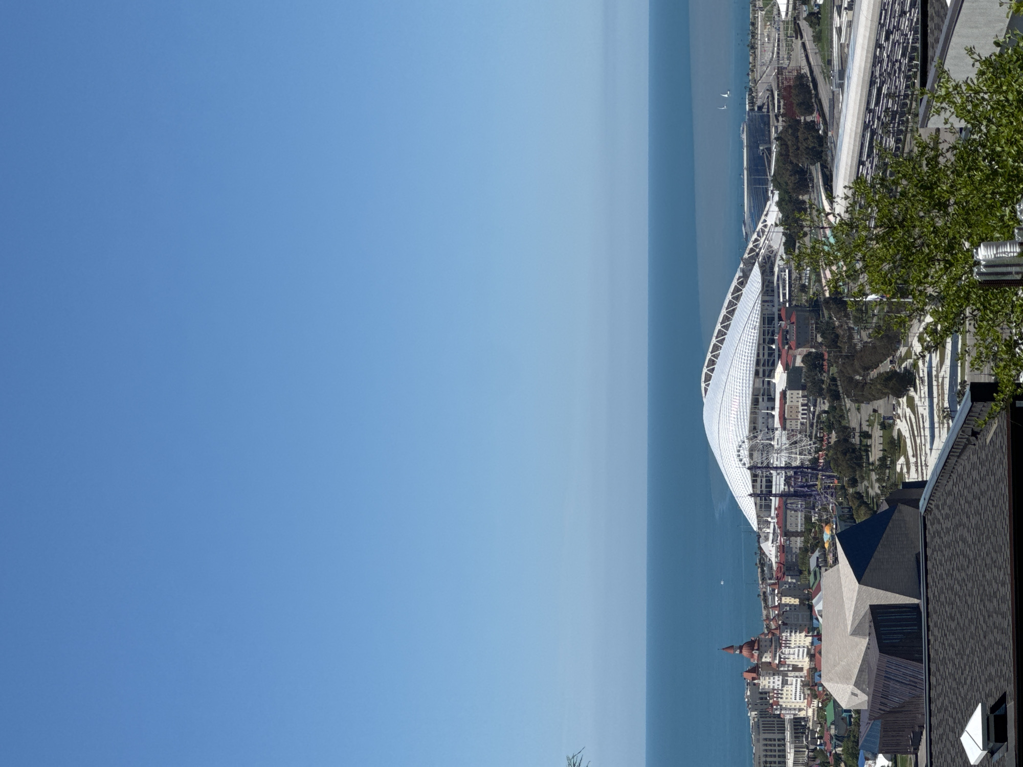 This high-angle, panoramic image captures a vibrant coastal cityscape in Sochi, Russia, under a clear and brilliant blue sky. The left side of the frame is dominated by the serene, expansive blue waters of what is likely the Black Sea, with two small boats visible in the distance.
The right side of the image showcases a dense urban and resort environment. A prominent feature in the mid-ground is a large, modern white building with a distinctive curved and patterned roof, identifiable as the Fisht Olympic Stadium. Adjacent to this stadium, an amusement park is visible, featuring a towering Ferris wheel and various other rides, indicating a lively recreational area.
Further inland, a multitude of buildings with diverse architectural styles, including some with traditional red-tiled roofs and others with contemporary designs, form the city's fabric. Green spaces, such as trees and landscaped areas, are interspersed among the structures. In the foreground on the right, a portion of a building with a dark grey, patterned roof and angular elements is visible, along with some green foliage, suggesting the photograph was taken from an elevated perspective overlooking the scene.
The image depicts a bright and sunny day, with no clouds in the sky, and the water appears calm. While no specific activities or individuals are clearly discernible, the presence of a stadium and amusement park suggests the location is a hub for leisure, tourism, and potential events.