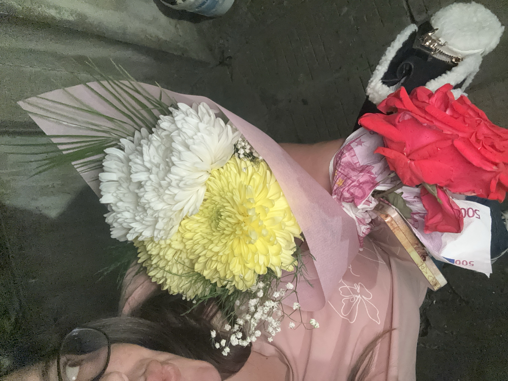 The image shows a view looking down, presumably from the perspective of a person holding two bouquets of flowers and some currency. The first bouquet consists of white chrysanthemums and yellow dahlias, wrapped in pink paper. The second bouquet includes red roses and appears to have multiple 500 Euro banknotes amongst the flowers. The person holding the bouquets is wearing glasses and a pink garment and has dark hair. In the background, parts of two pairs of shoes are visible. The environment appears to be outdoors with a dark, uneven surface. The location is specified as Razhevo Konare, Bulgaria, but there are no details in the image that confirm or deny this.