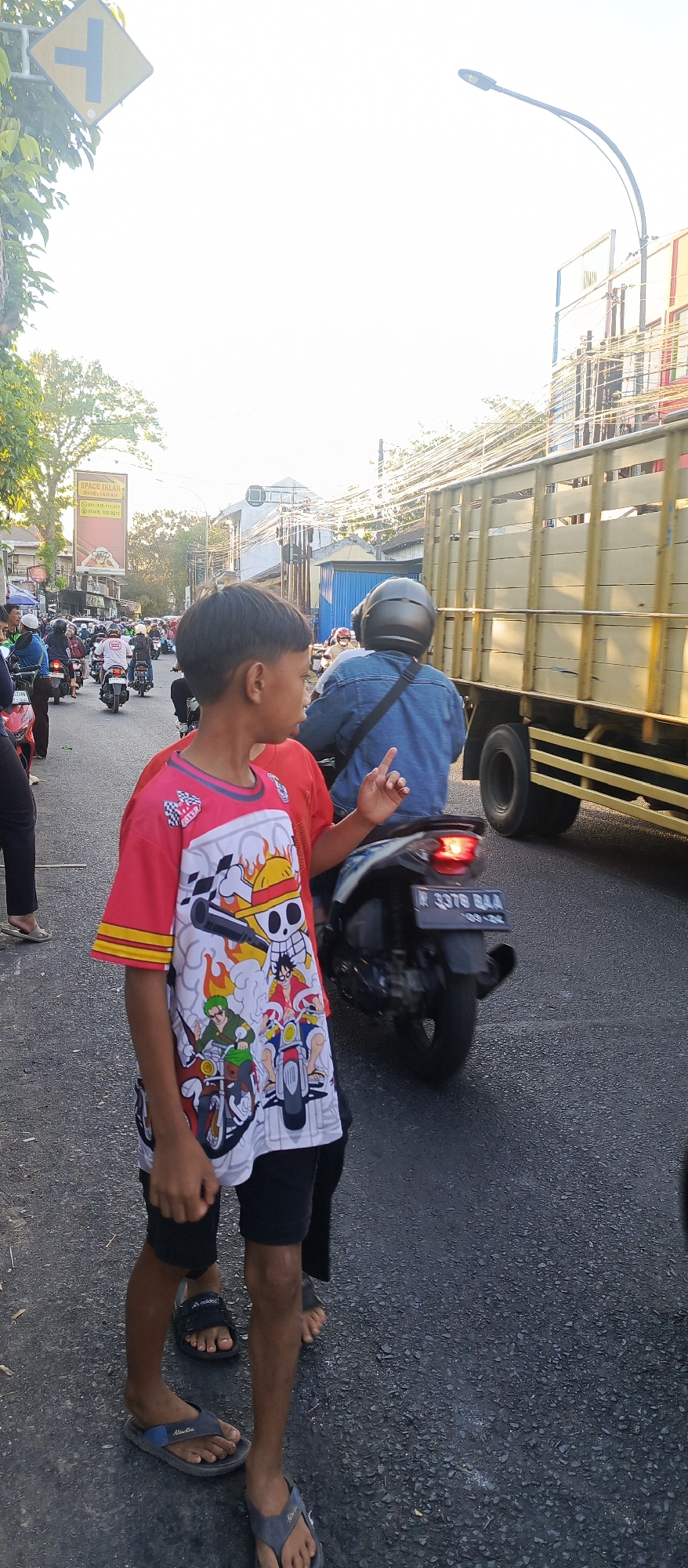 The image depicts a street scene in Malang, Indonesia, characterized by moderate traffic. A young boy in a red t-shirt adorned with anime characters stands in the foreground, facing away from the camera. He is wearing black shorts and sandals. Behind him, a person wearing a denim jacket and a black helmet rides a scooter. To the right, a yellow truck with a wooden cargo bed is moving along the road. Other vehicles and pedestrians are visible in the background, suggesting a busy urban environment. The sky is bright, indicating daytime. The road surface appears to be asphalt. A traffic sign is visible in the upper left corner of the image.