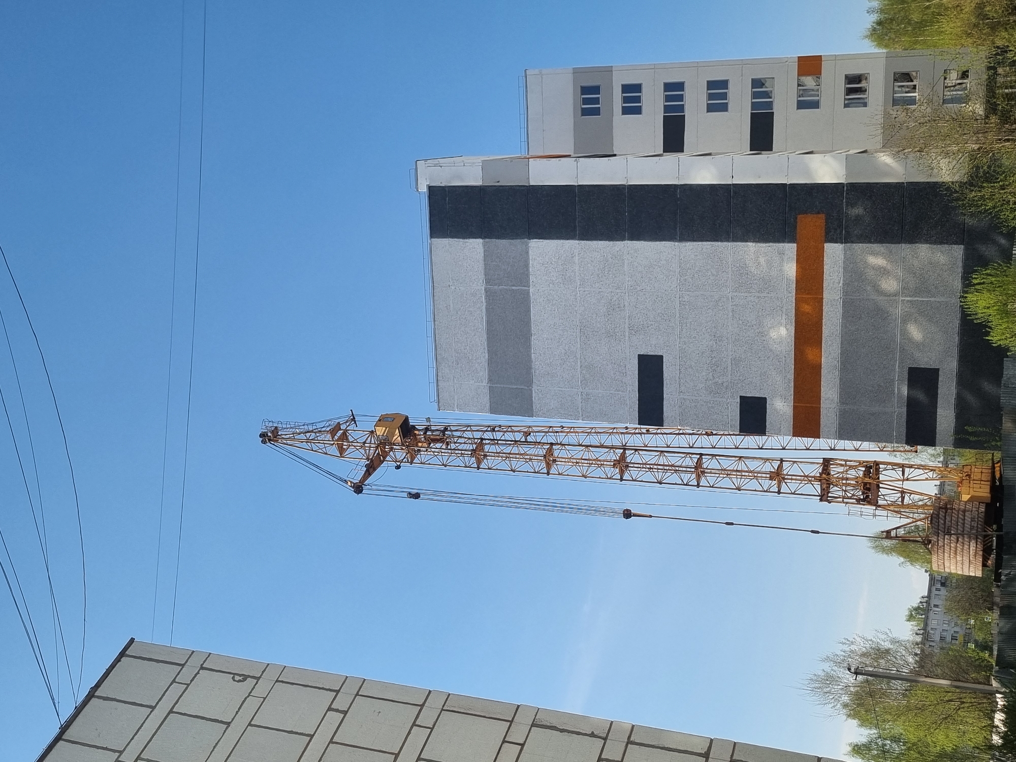 This image, taken from a low angle, showcases a scene in what is likely Chaikovsky, Russia, given the building style and environment. Dominating the view is a yellow construction crane with its boom extending diagonally across the frame against a clear blue sky. Power lines are visible to the left of the crane. The tops of two buildings are visible. The first building is grey. The second building is white. The second building has a dark grey band running horizontally near the top, with a vertical orange stripe on the right side. Beyond the buildings and crane, there are trees and rooftops of other buildings. The scene suggests an active construction area within the city.