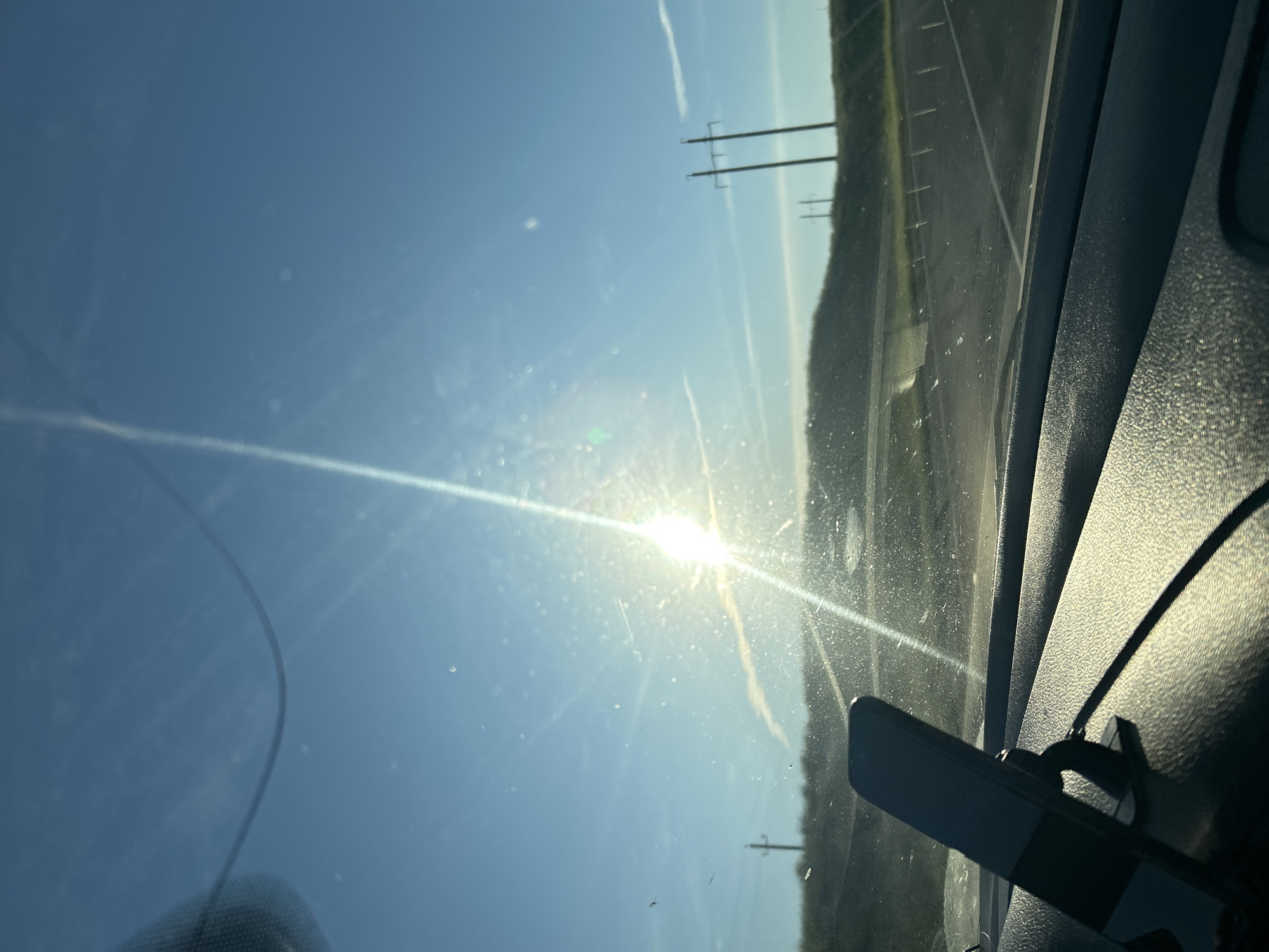 The image shows a view through a car windshield on a sunny day. The sun is bright and glinting off the windshield, creating lens flare. The sky is blue with some wispy clouds. In the distance, there are hills and power lines. The foreground shows the dashboard of the car, with a phone mounted on a stand. There is no indication of the location being Matrayevo, Russia.
