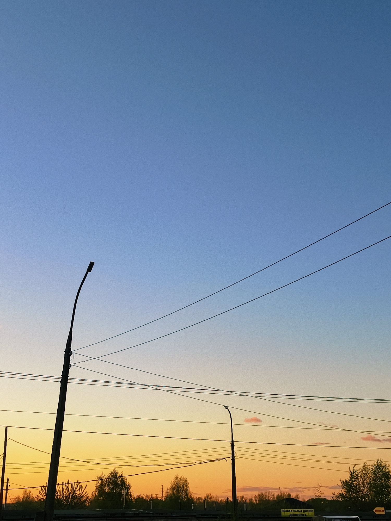 The image depicts a street scene in Chaikovsky, Russia at dusk or dawn. The sky transitions from blue at the top to orange near the horizon. Silhouetted against the skyline are trees, streetlights, and power lines. A sign with Russian text is visible.