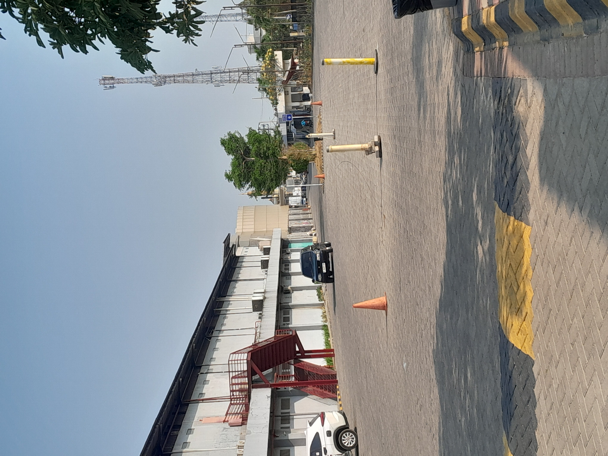 The image depicts an outdoor scene in Sempusari, Indonesia, featuring a long, grey building with a red exterior staircase. In the foreground, a paved lot is visible with yellow and black painted lines and several traffic cones. A black SUV is parked in front of the building, and a white SUV is to its left. Beyond the building, a telecommunications tower stands tall, and the sky is clear blue. The overall environment suggests a commercial or industrial area.