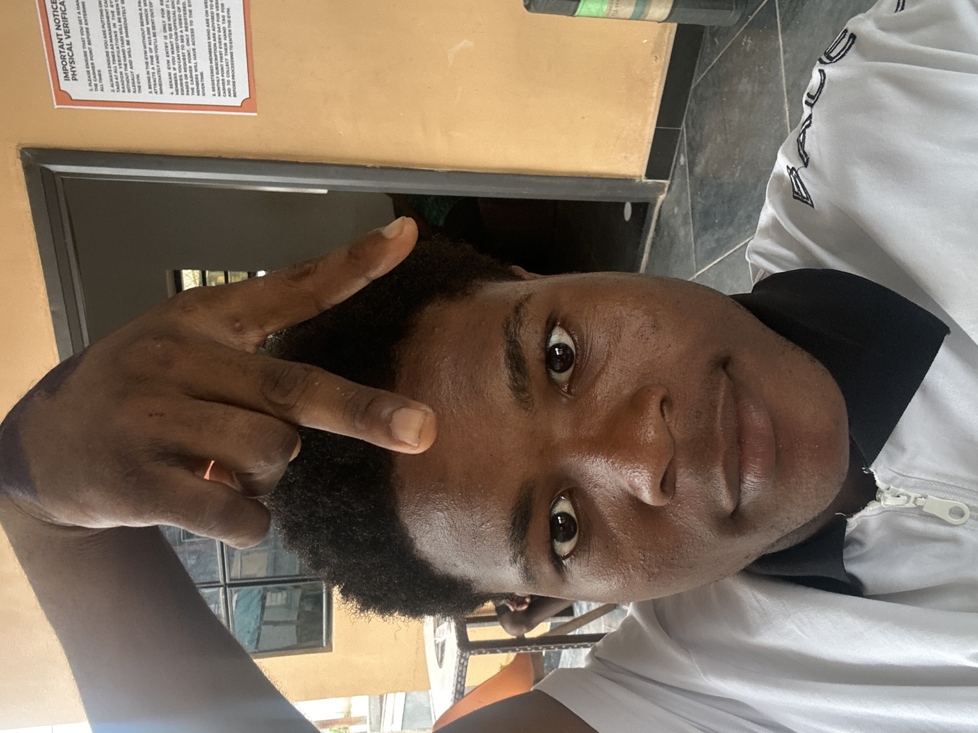 A close-up, front-facing selfie shows a young Black male in the foreground, with his right hand held above his head and bent at the wrist, with his index and middle fingers extended in a V-shape. He is wearing a white zippered jacket with a black collar and black trim on the shoulders with embroidered lettering. His hair is dark and curly. The background is slightly out of focus and shows an orange wall with a dark doorway. To the left, a small part of a metal table and chair is visible. A partially visible poster with text is on the orange wall to the right. The image appears to have been taken outdoors in a location that could be a residential or commercial area in Ihiagwa, Nigeria.