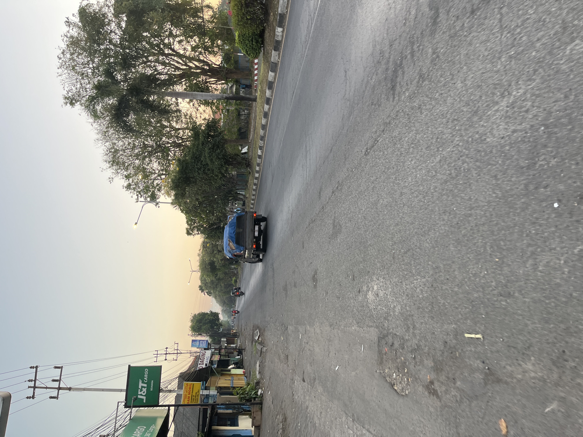 The image depicts a street scene in Ngaban, Indonesia, during what appears to be early morning or late afternoon due to the soft, warm light. The road is paved and shows some wear and tear, with a few potholes visible on the left side. Along the left side of the street, there are buildings, some with storefronts, and overhead power lines supported by utility poles. Signs are visible on the buildings, including one for "J&T CARGO" and another for "SAM MOBIL". Several motorcycles and a small truck are present on the road. The truck in the foreground is carrying a load covered by a blue tarp. Trees line the right side of the road, with lush foliage and a palm tree prominently featured. A sidewalk with a concrete curb, painted black and white, runs alongside the buildings and trees. The sky is clear with a gradient of light colors, suggesting diffused sunlight. The overall atmosphere is calm and suggests a typical urban or semi-urban environment in Indonesia.