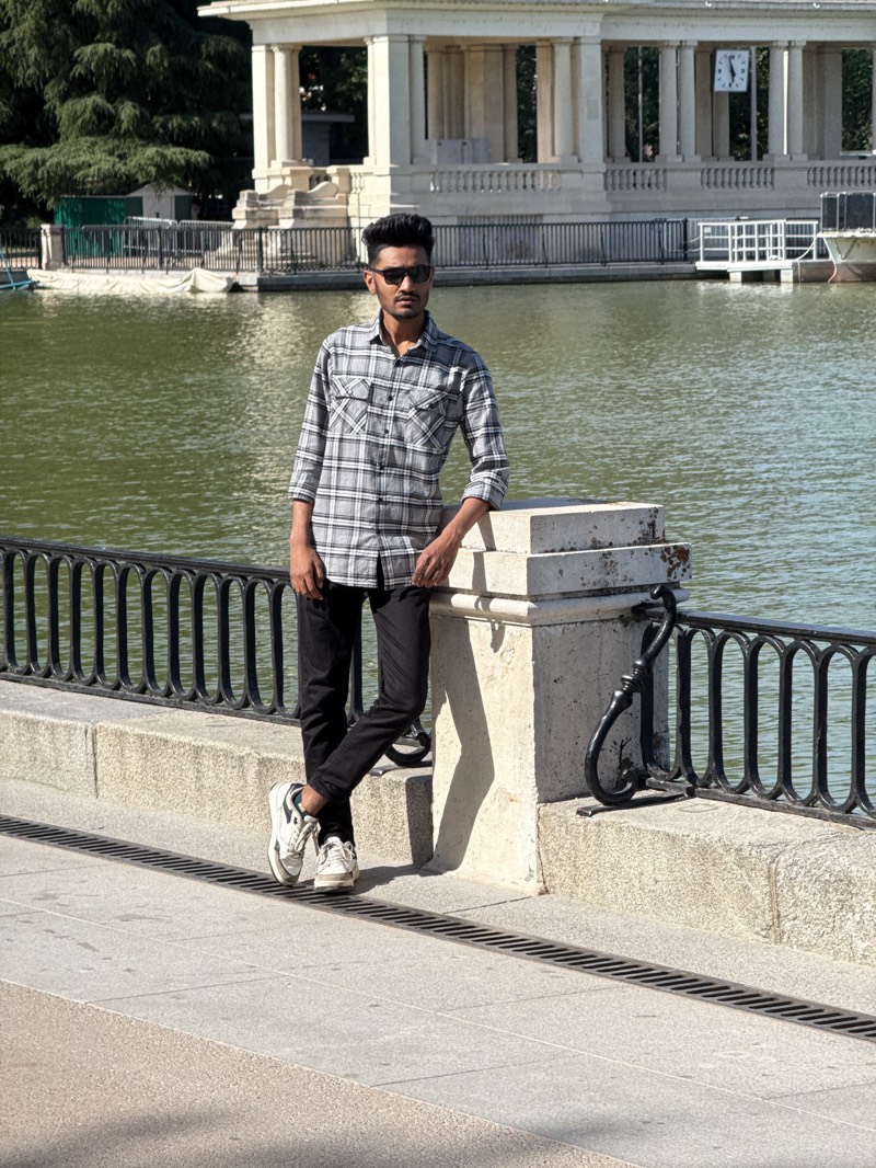 A young man stands casually by the lake in El Retiro Park, Madrid, Spain, posing for a photograph. He is positioned in the center of the frame, leaning against a light-colored stone pillar that forms part of a black metal railing, which separates the paved walkway from the water.
The man, likely in his early twenties, has dark hair styled upwards and wears dark sunglasses. He is dressed in a grey, white, and black plaid long-sleeved shirt, black trousers, and white sneakers with black accents. His stance is relaxed, with one foot casually crossed over the other and his forearms resting on the pillar.
Across the greenish water of the lake, in the background, stands a grand, classical-style white stone monument with numerous columns, strongly resembling the Monument to Alfonso XII. A clock face is visible on the upper left section of this structure, with hands indicating approximately 12:00. Several small boats, some covered with white tarps, are docked near the monument, suggesting boat rental services. Lush green trees are visible behind and to the left of the monument.
The scene is brightly lit by natural sunlight, casting long shadows from the man and the pillar onto the light-colored paved walkway, which features a metal drain grate running across it. This indicates a clear, sunny day, likely during late morning or early afternoon. The man's expression, partially obscured by sunglasses, appears neutral or contemplative.