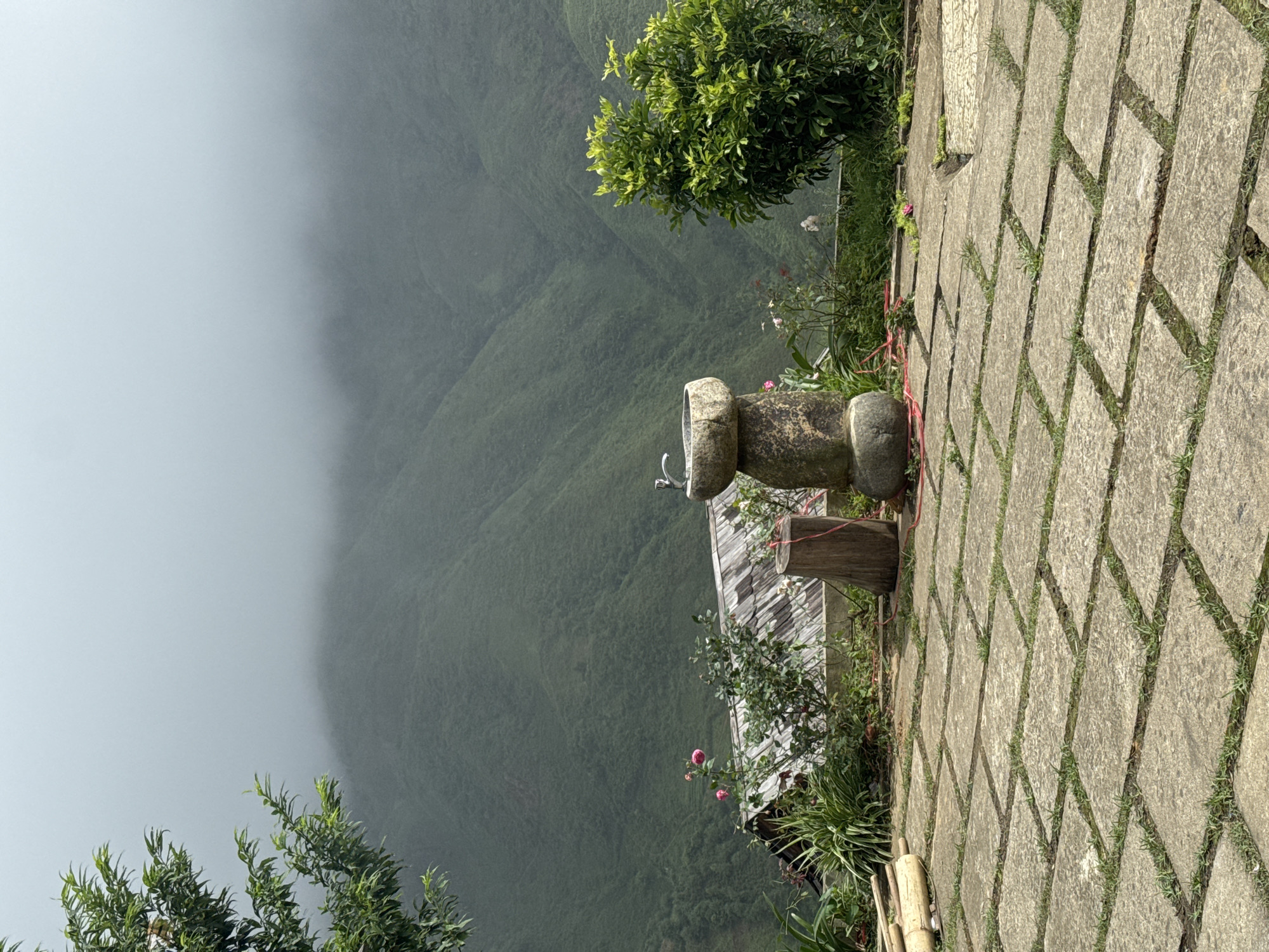This is a medium shot, exterior photograph of a mountain view from a paved stone patio. In the foreground, a paved stone patio is visible with grass growing between the stones. A tree with green leaves is on the left side of the frame. In the middle ground, there is a stone sink with a faucet, a wooden post, a small house with a corrugated metal roof, and a bush with green leaves on the right side. Pink flowers are also visible. In the background, lush green mountains are shrouded in fog. The scene appears to be a scenic overlook or a garden area in Ta Xua, Vietnam.