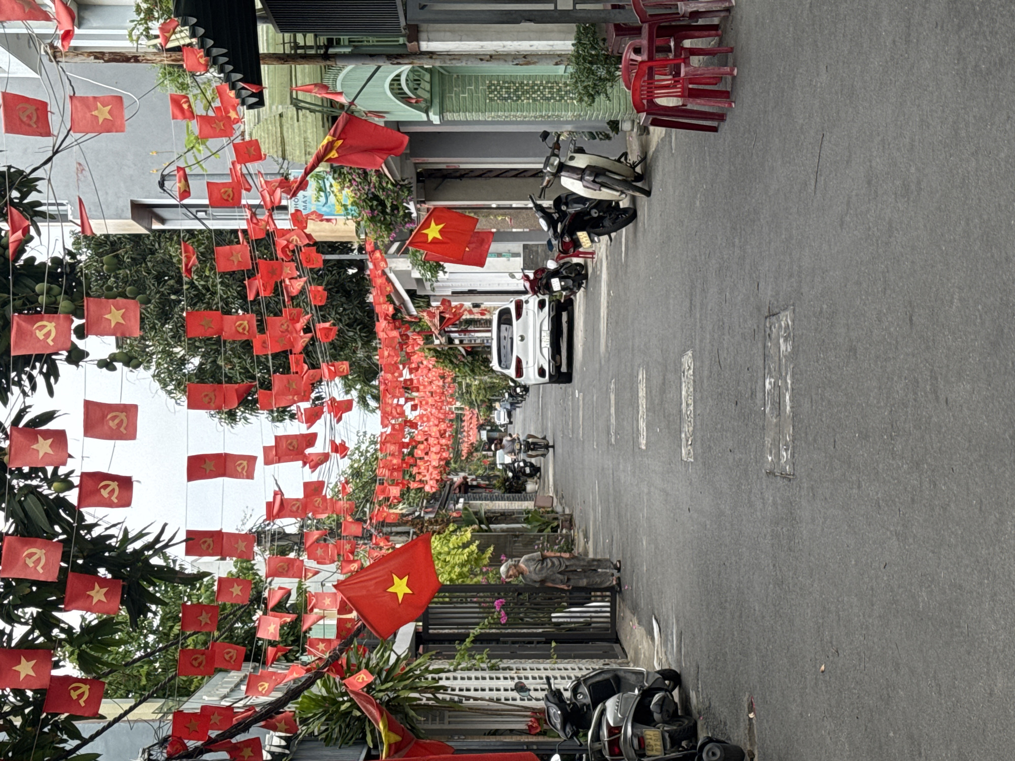 A straight, street-level shot shows a narrow street in Đà Nẵng, Vietnam, densely decorated with Vietnamese flags hanging overhead. The street is paved with gray asphalt, displaying occasional white paint markings. Along both sides of the street, buildings are visible, constructed in a variety of styles and colors ranging from gray to green. Motorbikes are parked intermittently along the curbs, while a white car is parked further down the street. A person is walking alongside a building. The image is taken during daylight hours.