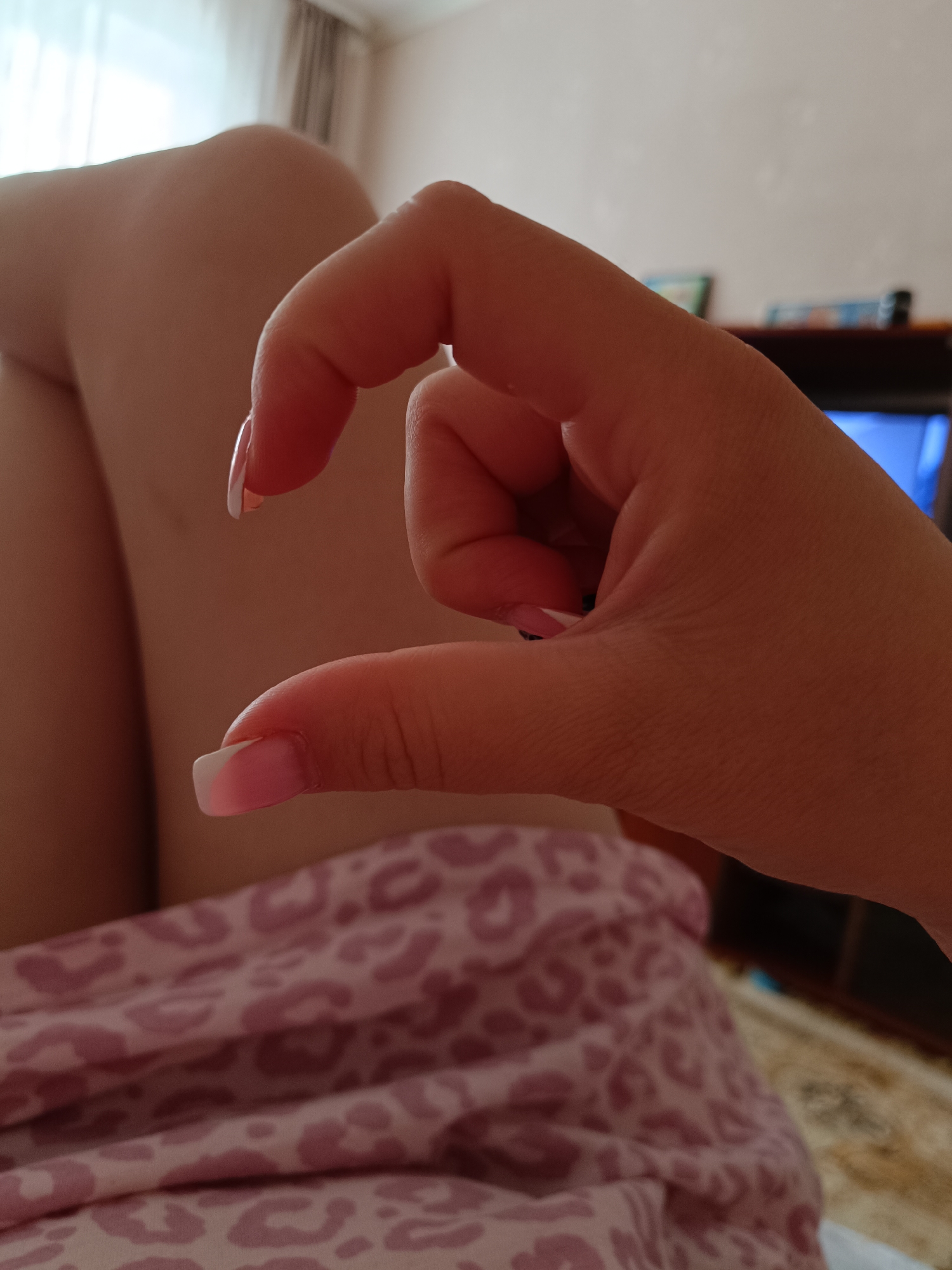 A human hand with manicured fingernails forms a small heart shape. The hand is positioned in the foreground, partially obscuring the view behind. The subject is sitting, and the legs are visible. The background includes a leopard print patterned garment and a television in the background. The location appears to be indoors. I cannot verify the image location in Tobolsk, Russia.