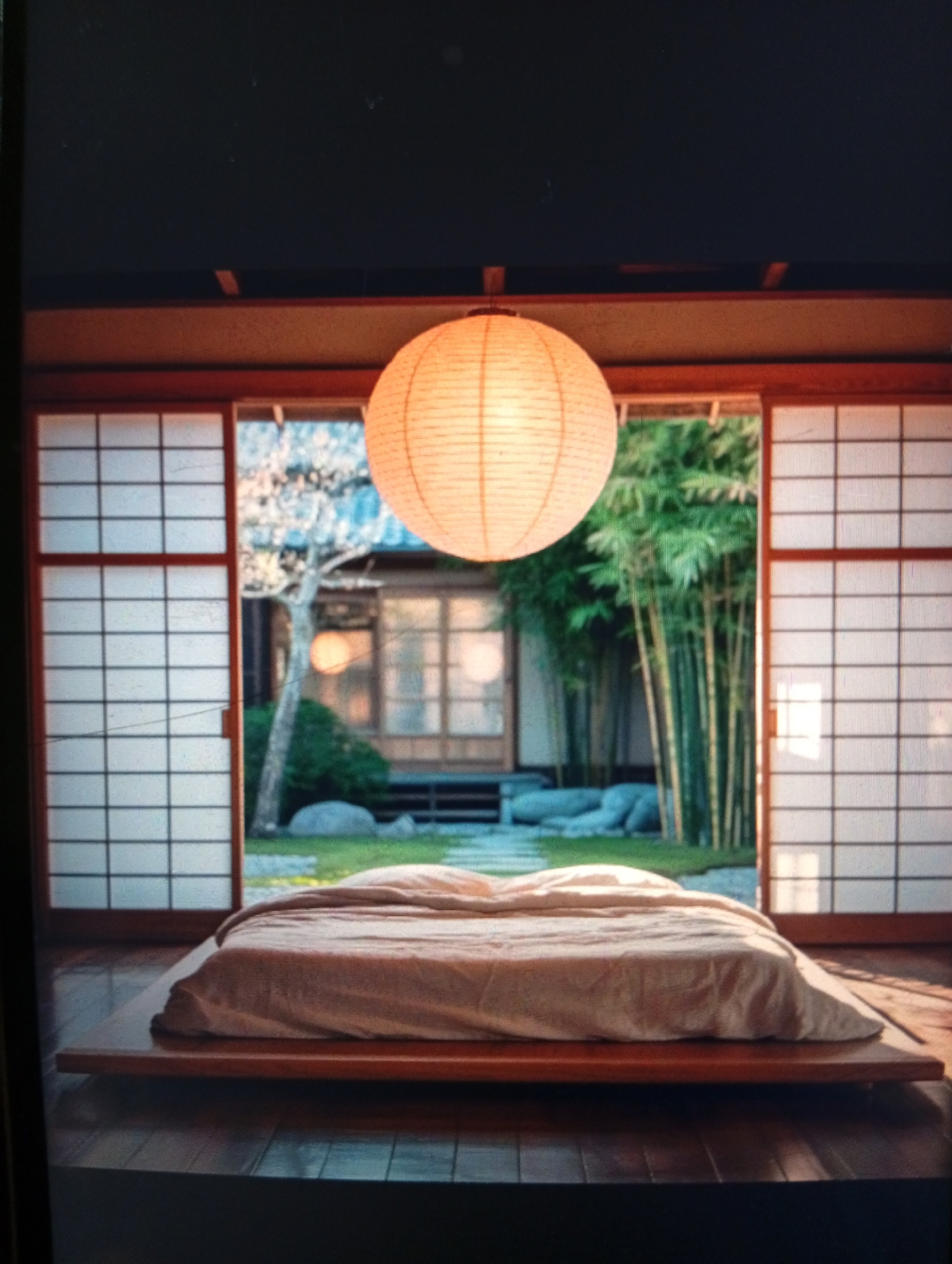 A room with a Japanese aesthetic features a low platform bed centered beneath a paper lantern. Sliding doors with a grid pattern partially obscure a garden view including bamboo trees and decorative rocks. There are no people present in the image. The location is not in the city of Dutsen Alhaji, Nigeria.