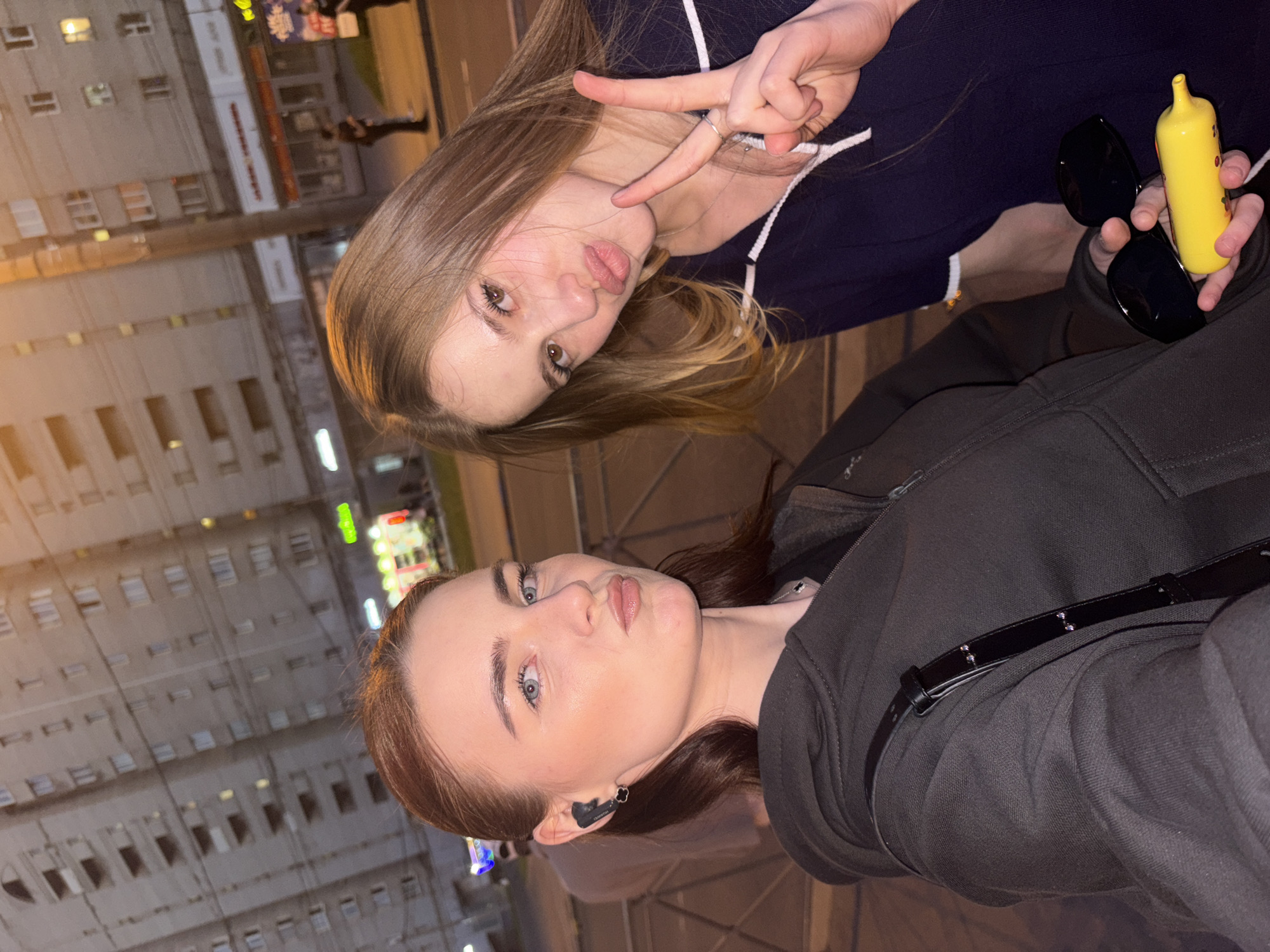 Two young adult women are captured in a close-up, chest-up selfie format, positioned in an urban environment at twilight or night.
The woman on the left, with dark brown hair pulled back, gazes directly at the camera with blue eyes and a slight pout. She wears a dark grey or black zip-up hoodie and a black wireless earbud in her right ear (viewer's left). A thin silver chain is visible around her neck, and a black strap, likely from a bag, crosses her shoulder.
The woman on the right, with long light brown hair parted centrally, also looks at the camera with brown eyes and a pout. Her left hand is raised, forming a "peace" sign with her index and middle fingers. She wears a dark blue polo shirt with a white collar and white stripe detailing on the sleeves. In her right hand (viewer's left), she holds a pair of black sunglasses and a bright yellow, elongated, squeezable toy or novelty bottle with red accents.
The background features multi-story residential or mixed-use buildings, characteristic of Soviet-era architecture, with light grey or beige facades and numerous rectangular windows, some illuminated. At street level, various commercial storefronts are visible with glowing signs, including a prominent green neon sign on the far left resembling "Ozon" (a major Russian e-commerce company). Streetlights and utility poles are present. Blurred figures of other pedestrians can be seen further down the street. Metal crowd barriers are in the immediate foreground, positioned between the subjects and the distant background. The overall lighting is artificial, emanating from streetlights and shop windows, suggesting a late evening or nighttime scene.
The architectural style of the background buildings and the identifiable Russian retail presence (Ozon) are highly consistent with urban environments in Saint Petersburg, Russia, though a precise street name cannot be identified solely from this image.
