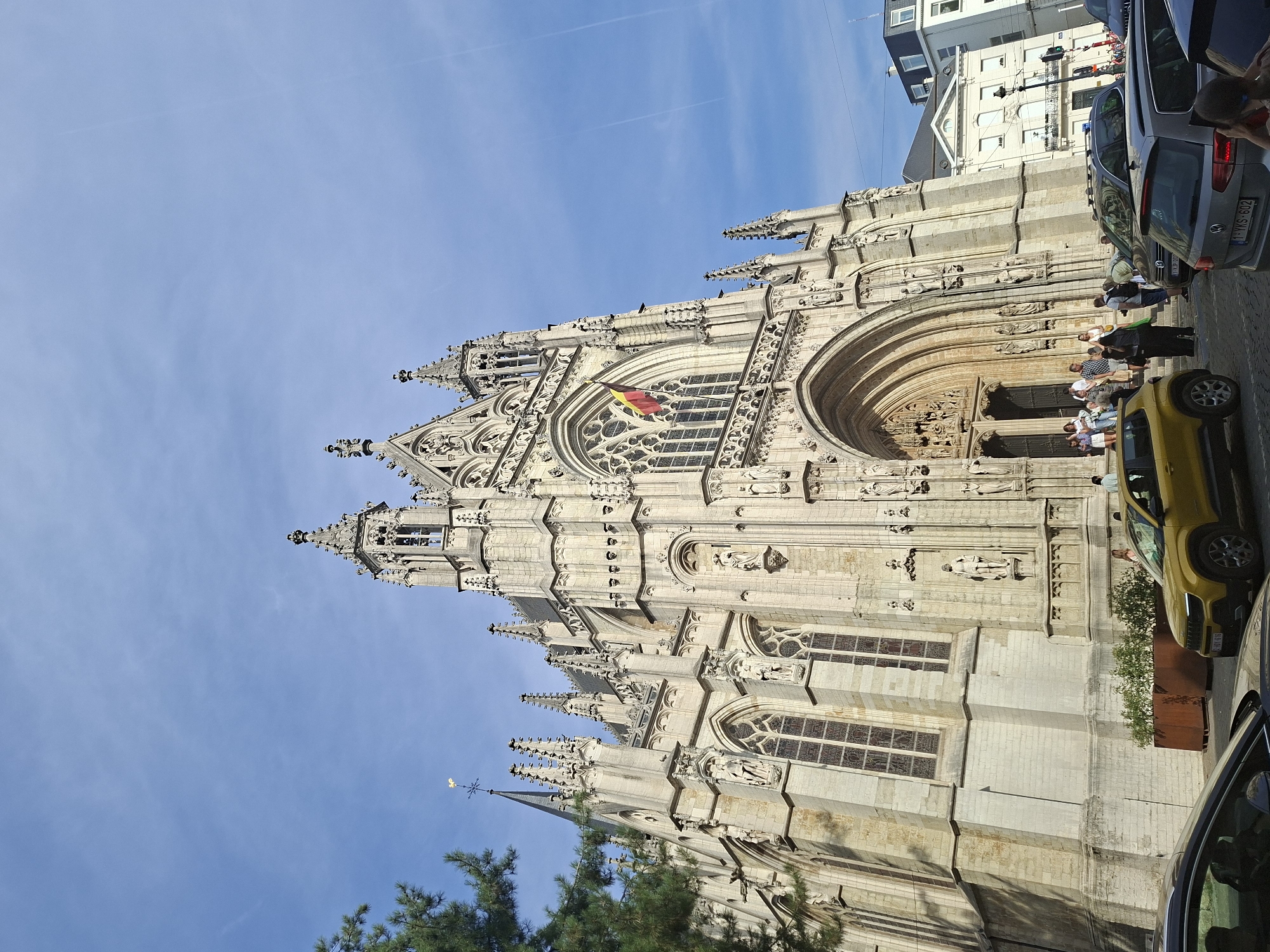 The image shows the exterior of the Notre-Dame du Sablon church in Brussels, Belgium, on a sunny day. The gothic architecture of the church is prominent, with intricate stonework, tall spires, and arched windows. A Belgian flag is visible hanging from a window on the facade. In front of the church, there is a street with several cars parked, including a yellow SUV and a silver hatchback. A small group of people are gathered near the entrance of the church, some appearing to be tourists. Green trees are visible on the left side of the frame, and the sky is clear blue with some faint contrails. The overall scene depicts a bustling urban environment with a significant historical landmark.