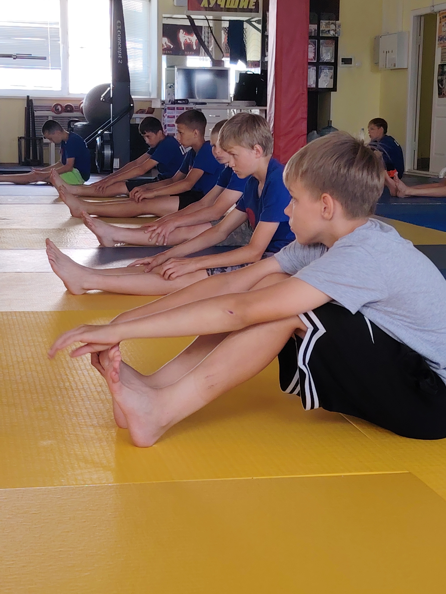 This image depicts a group of young boys in athletic attire engaged in a stretching exercise on a yellow mat, likely in a gymnasium or training facility. The boys are seated with their legs extended forward, reaching for their toes, in a common pre-exercise warm-up. They appear to be of similar age, possibly pre-teens or early teenagers. One boy in the foreground is wearing a grey t-shirt and black shorts with white stripes. Other boys are seen in blue t-shirts. In the background, there are rowing machines, weightlifting equipment, and posters on the wall, suggesting a sports training environment. The room has bright lighting, with windows visible on the left side of the image. While the specific location within Chaikovsky, Russia, cannot be precisely identified from the image alone, the presence of sports equipment and training activities strongly suggests a connection to a local sports club, school gymnasium, or a dedicated martial arts/fitness center.