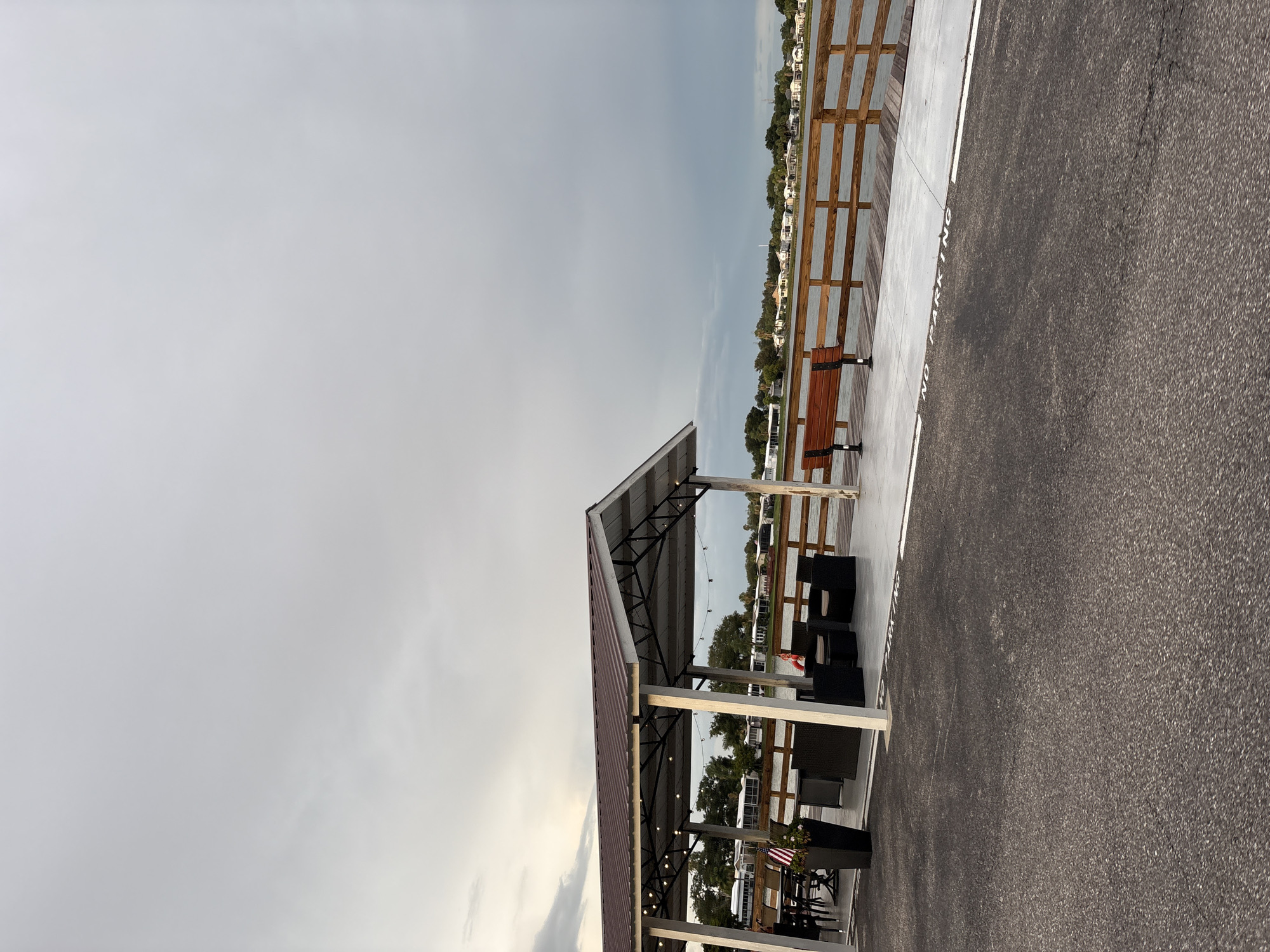 The image depicts an outdoor seating area on a pier, located in Bradenton, United States. The pier features a covered structure with a metal roof, supported by white pillars, and strung with decorative lights. Underneath the shelter, there are several pieces of dark-colored outdoor furniture, including chairs and a table, arranged for seating. Adjacent to the seating area, a wooden bench is positioned on the pier. A wooden railing runs along the edge of the pier, overlooking a body of water. In the background, across the water, a line of houses and trees is visible. The sky is overcast with gray clouds. In the foreground, a paved area with "NO PARKING" painted in white lettering is visible.