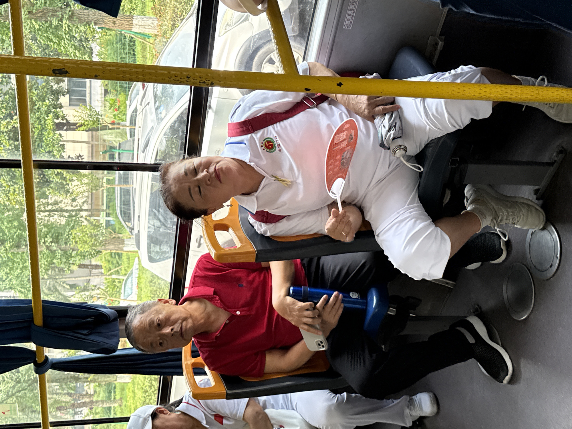 The image captures the interior of a public bus in Jiangjin, China, providing a glimpse of passengers and the urban environment outside.
In the mid-ground, an elderly Asian man is seated on the left, wearing a red polo shirt, dark trousers, and black and white athletic shoes. He possesses short, greying hair and is looking towards the left out of frame. His left hand holds a light-colored smartphone with a triple-lens camera module, while his right hand rests on a blue thermos bottle positioned between his knees.
Adjacent to him on the right, an elderly Asian woman is seated, adorned with small white hoop earrings and dark hair pulled back. She wears a white polo shirt featuring a circular red and green logo with the Chinese characters "中国力量" (China Power), white capri pants, and light-colored sneakers. Red backpack straps are visible over her shoulders. She holds a red, circular fan in her right hand and has a folded grey umbrella or fabric item resting on her lap. Her eyes are partially closed, and her facial expression suggests weariness or concentration while fanning herself.
A third passenger, partially visible on the far left, wears a white cap, a white polo shirt, and white pants.
The bus interior features orange-backed seats with dark blue cushions. Prominent yellow vertical and horizontal grab poles traverse the space. A dark blue fabric, possibly a curtain or shade, is tied around the horizontal pole above the man. The floor is grey, marked with two circular silver-colored access covers. A white sticker with Chinese text is affixed to the window frame on the right.
Through the large windows, an exterior view reveals abundant green trees and bushes, indicative of a verdant urban or suburban landscape. Several vehicles, including a light-colored SUV, are visible on what appears to be a street or parking area. Distant buildings are discernible amidst the foliage.