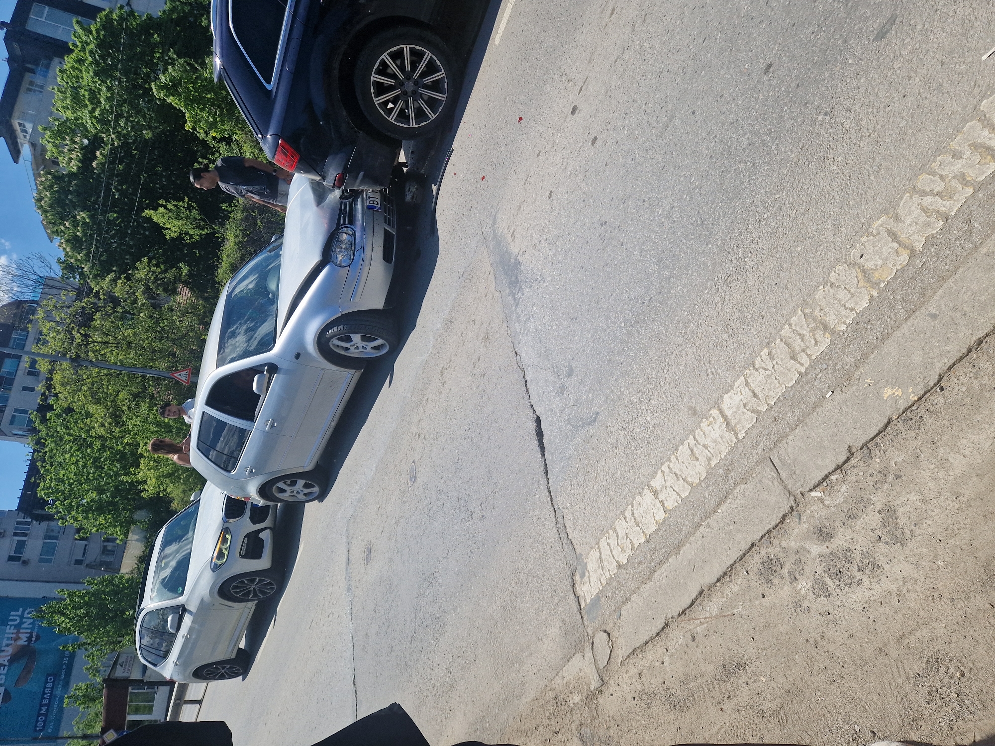 Here is a description of the image:
The image appears to depict a traffic incident on a city street. A silver Volkswagen Golf has collided with a dark-colored SUV in front of it. There's damage to the front of the Golf and the rear of the SUV. People can be seen near the vehicles, possibly assessing the situation or involved in the incident. The accident seems to have occurred on a paved street with a white lane divider. In the background, there are buildings, trees, and advertisements including a billboard. One can spot the license plate on the Golf starts with "B1".
