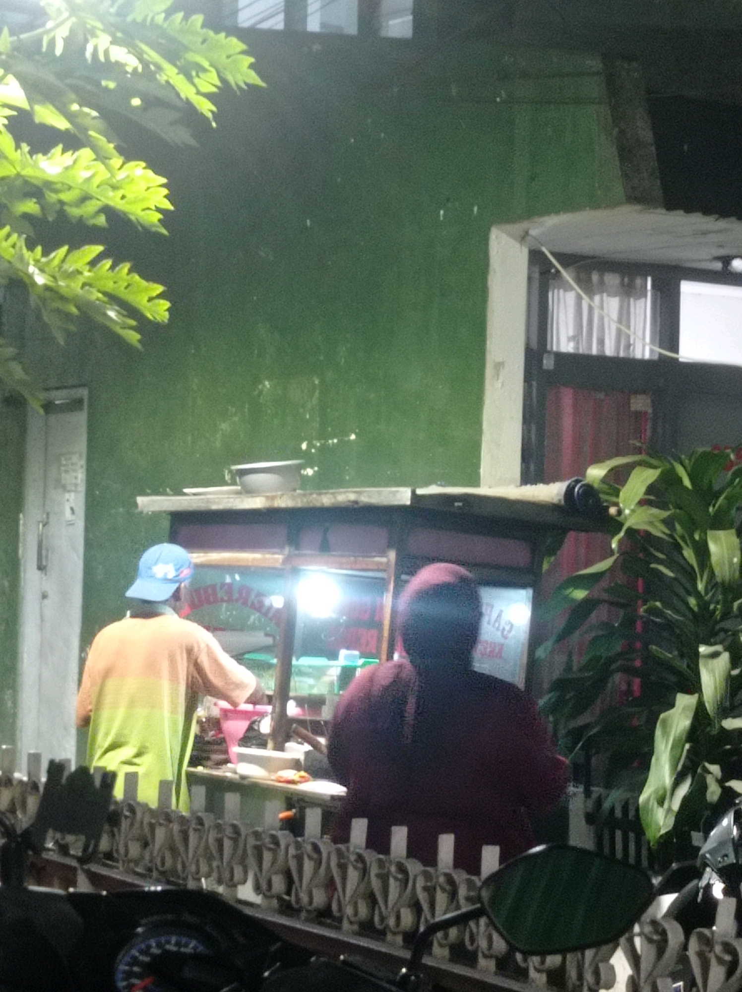 A nighttime scene in Bandung City, Indonesia, shows two individuals standing near a food stall. A person wearing a blue baseball cap and an orange and green gradient shirt stands facing a food cart. Their back is to the camera. To their right, another person, wearing a dark maroon top, is also facing the food cart. The food stall is illuminated by bright lights, highlighting various items inside. In the foreground, a fence runs horizontally across the frame, with a motorcycle visible to the left. Lush green foliage is present on the left side of the image, extending from the upper left corner. The background features a textured green wall and architectural elements, including a window with curtains and a doorway on the left.