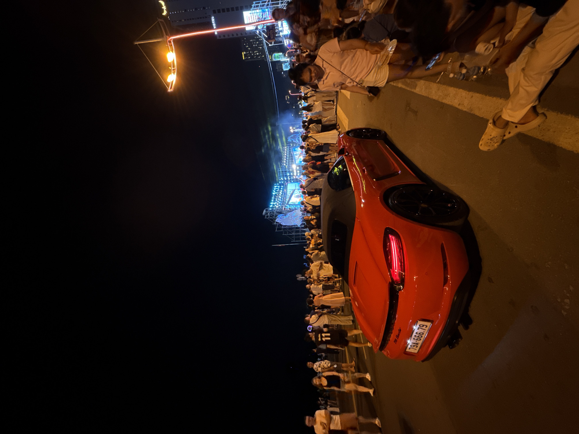 A red Porsche convertible is parked on a street at night, its license plate visible as 79A-666.79. The car is positioned on the right side of the frame, facing away from the viewer and slightly to the left. In the background, a crowd of people are gathered on the sidewalk, facing a large stage setup with bright lights and structures that suggest an outdoor event or concert. The event appears to be taking place along a coastal area, with tall buildings visible in the distance, including one labeled "NOVOTEL." The scene is illuminated by streetlights and the lights from the stage. The image captures a lively atmosphere in a city setting, likely during a nighttime festival or event in Nha Trang, Vietnam.