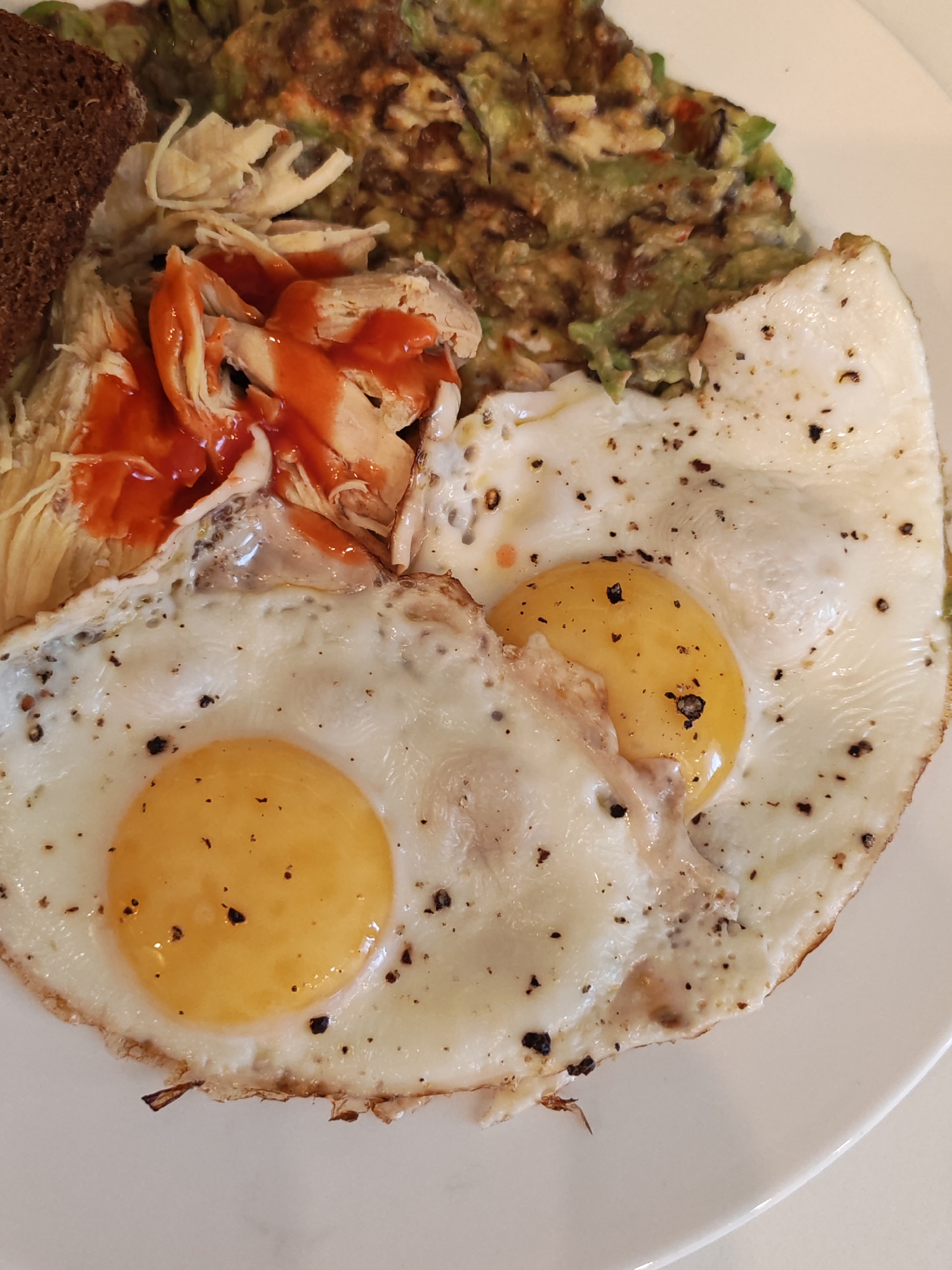 A close-up, overhead shot displays a meal on a white plate. The dish features two fried eggs with visible black pepper and seasoning. To the left of the eggs is a pile of shredded chicken drizzled with a red sauce, and a slice of dark bread. Behind the chicken and to the right of the eggs is a portion of what appears to be avocado or guacamole, mixed with other ingredients. The lighting is natural, and the focus is sharp, highlighting the textures and details of the food. The meal is served in an indoor setting, likely a home or restaurant. The location is Unknown, Unknown.