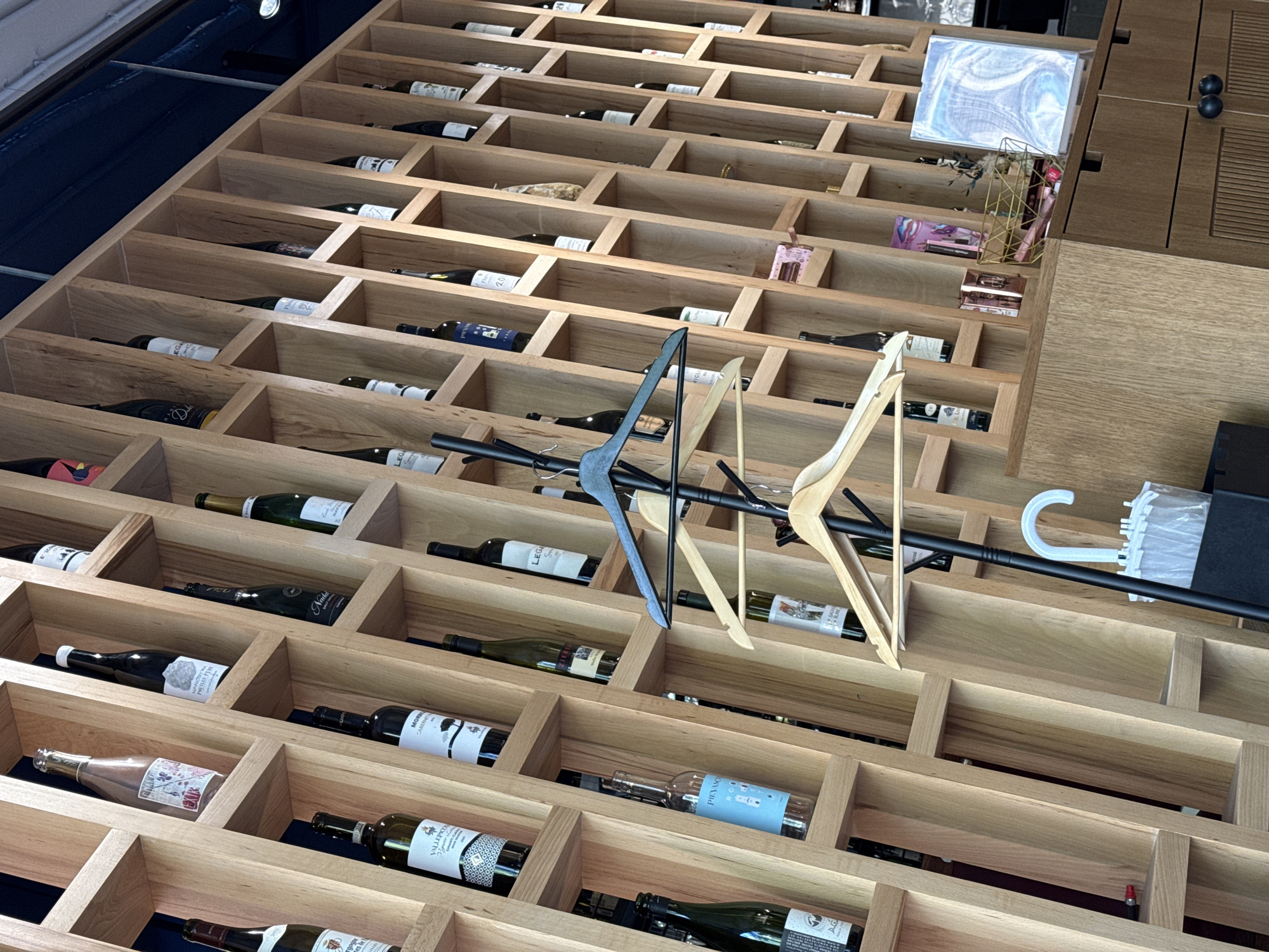 A slightly elevated, oblique view captures an intricate wooden shelving unit designed for wine storage or display. The unit, made of light-colored wood, features numerous rectangular cubbies, each housing a wine bottle laid horizontally. A dark blue surface is visible along the upper left edge of the image, suggesting a deeper recess or a wall behind the shelving.
Numerous wine bottles, mostly dark green or brown glass, are neatly arranged within the cubbies. Many have their labels facing upwards, displaying various designs and some discernible text. Visible brand names and varietals include "LEGA," "VALLARCO," "MORMONA," "PIEVASO" (on a bottle with a blue label), "CABERNET," and "PINOT NOIR." Some labels also show vintage years such as "2019" or "2020."
In the central area, several clothes hangers are prominently displayed, suspended from a black horizontal bar that runs across multiple cubbies. There are wooden hangers and a light blue plastic hanger. Further to the right, a white plastic hook, possibly part of a clothes hanger, is visible, along with a stack of white plastic items.
Towards the upper right, the shelving unit transitions into a flat wooden surface. On this surface, there's a white rectangular object with a swirling blue and purple pattern, alongside a gold-colored wire basket containing small pink items, possibly cosmetics or small decorative objects. The overall impression is an organized retail or display space, possibly a wine shop, cellar, or a section within a larger store that also sells other goods. The lighting is even, suggesting an indoor, artificial light source. No people are visible.