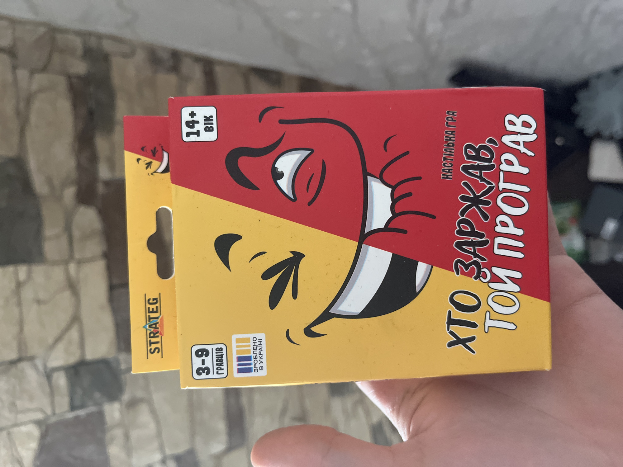 The image shows a close-up of a person's hand holding a game box. The game box is predominantly yellow and red, with cartoon-like faces on its front. The text on the box is in Ukrainian. In the top left corner, the word "STRATEG" is visible. Below this, a box indicates "3-9 гравців" (3-9 players). Further down on the yellow side, there is text that appears to say "ЗРОБЛЕНО В УКРАЇНІ" (Made in Ukraine) next to a barcode-like image. The right side of the box is red and has a white square with "14+ ВІК" (14+ age) inside. The main text on the front of the box reads "ХТО ЗАРЖАВ, ТОЙ ПРОГРАВ" (He who laughed, lost). The background appears to be a textured wall. The location is not specified in the image or the provided context.