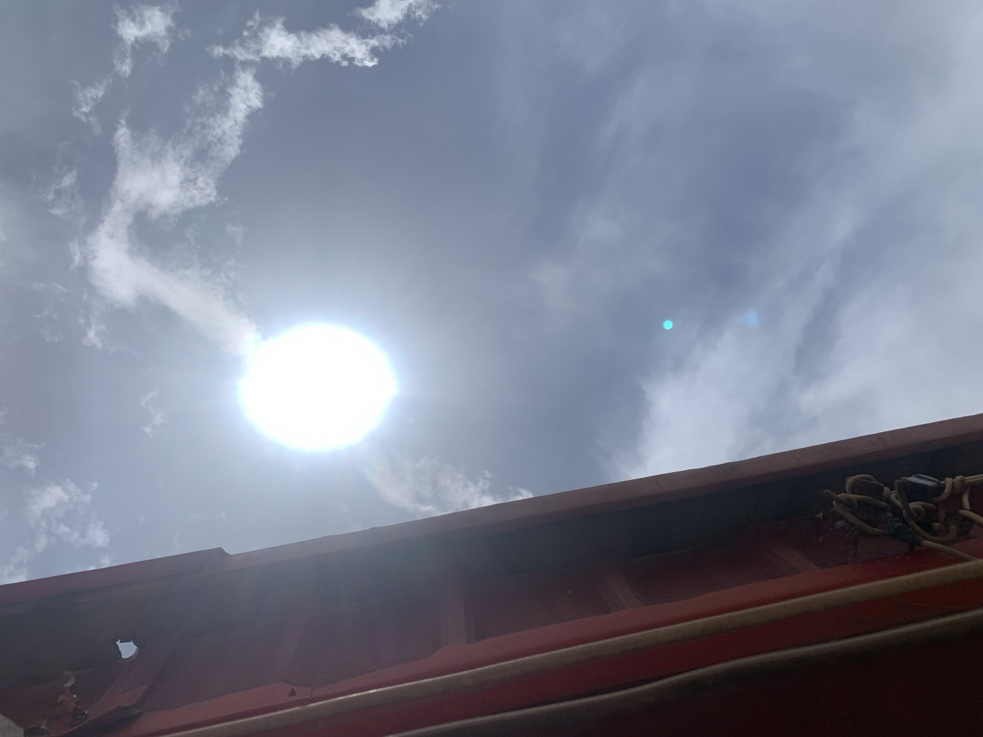 The image depicts a view upwards towards a bright sun in a sky with scattered clouds. A section of what appears to be a red-painted metal roofline occupies the lower portion of the frame. Some cables or ropes are visible along the edge of the roof. The location is Barakin Akawo, Nigeria.
