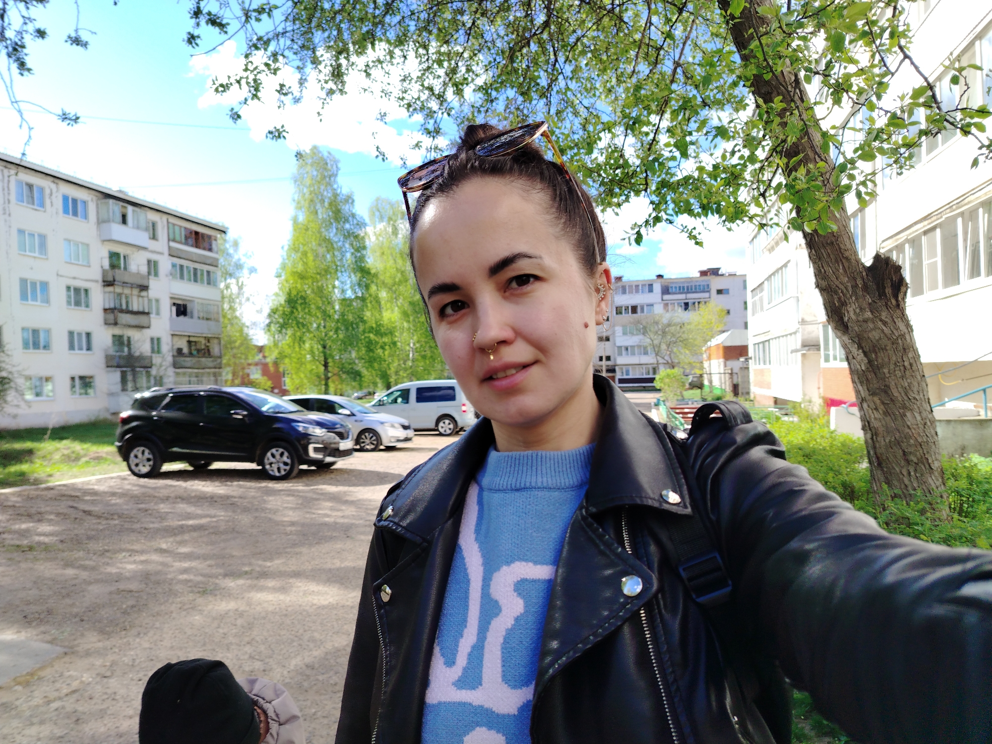 A woman is taking a selfie outdoors in Noviy Urengoy, Russia. She is wearing a blue sweater with a white pattern, a black leather jacket, and has sunglasses perched on her head. Buildings are visible in the background, along with several parked cars, including a black SUV. A tree is to her right, framing part of the image. The ground appears to be a paved surface. The sky is visible through the leaves of trees overhead, suggesting a clear day.