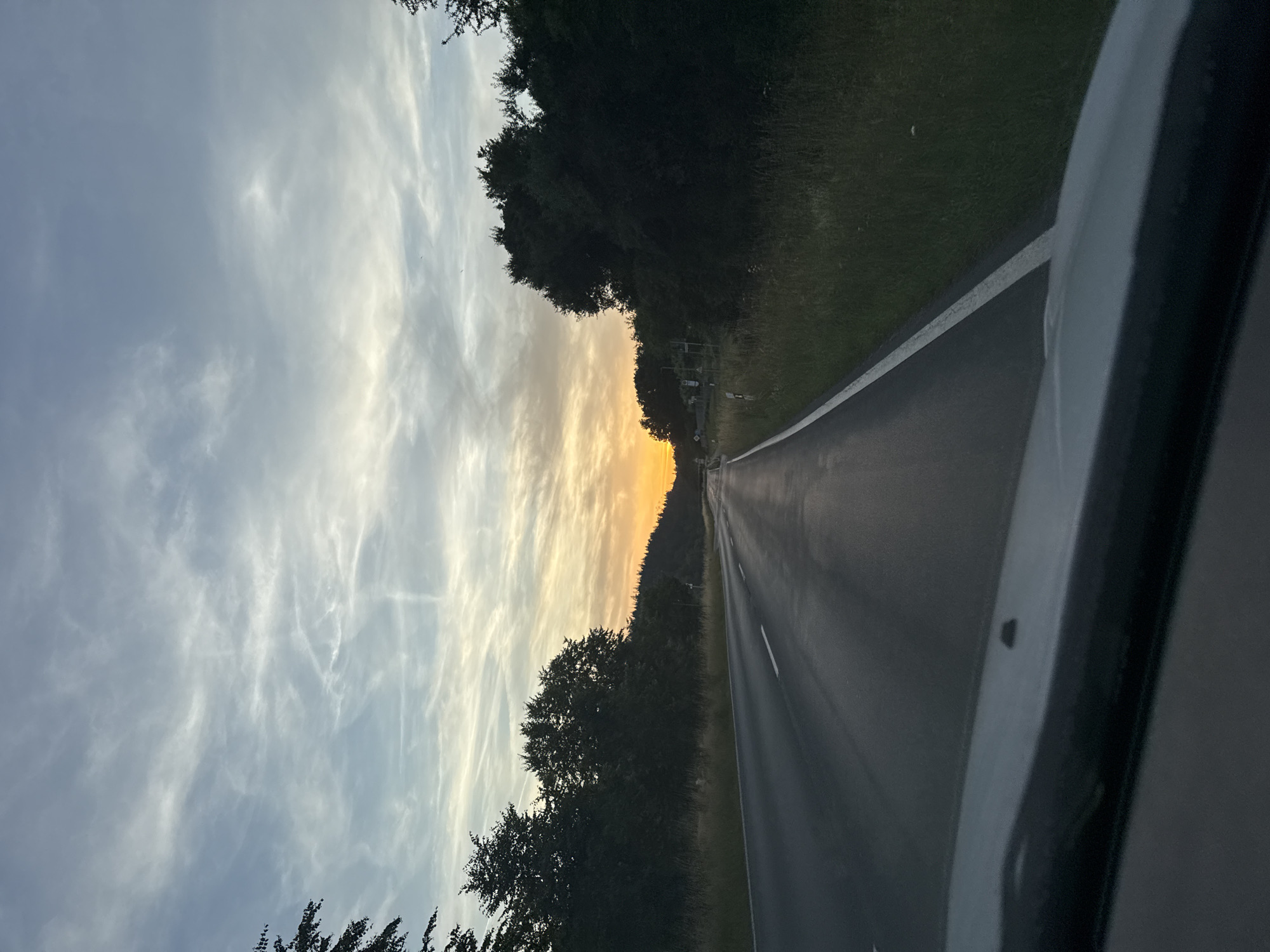 This is a vertical, eye-level shot of a road curving through a wooded area at sunset. The road is asphalt with a white line marking the center. On the left side of the road, there are dense trees, and to the left of the trees, a vast expanse of sky. The sky is filled with wispy clouds, tinged with the warm hues of orange and yellow from the setting sun. The right side of the image shows the car's interior, a dark, rounded frame, suggesting the photo was taken from inside a vehicle. The scene evokes a sense of journey and the peaceful beauty of nature at dusk. The location is identified as Brandoberndorf, Germany. No people or text are visible in the image.