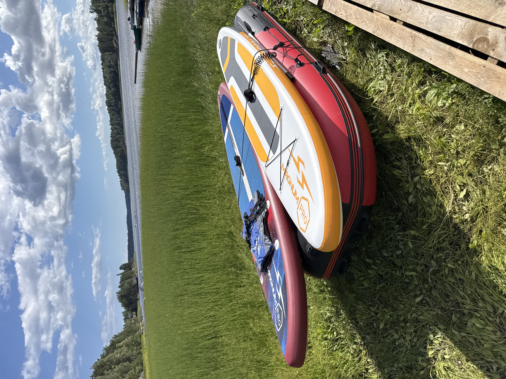 In the foreground of this image, three inflatable watercraft are resting on a grassy bank beside a body of water. The topmost item is a red inflatable boat, and stacked on top of it are two stand-up paddleboards. The bottom paddleboard is blue with red and white accents, and a blue life jacket is draped over it. The top paddleboard is white with orange and grey stripes and a black coiled leash. The background features a wide expanse of calm water, with a tree-lined shore visible in the distance under a bright blue sky dotted with fluffy white clouds. The environment suggests a recreational setting, likely near Lumivaara, Russia, given the presence of water and natural landscape.