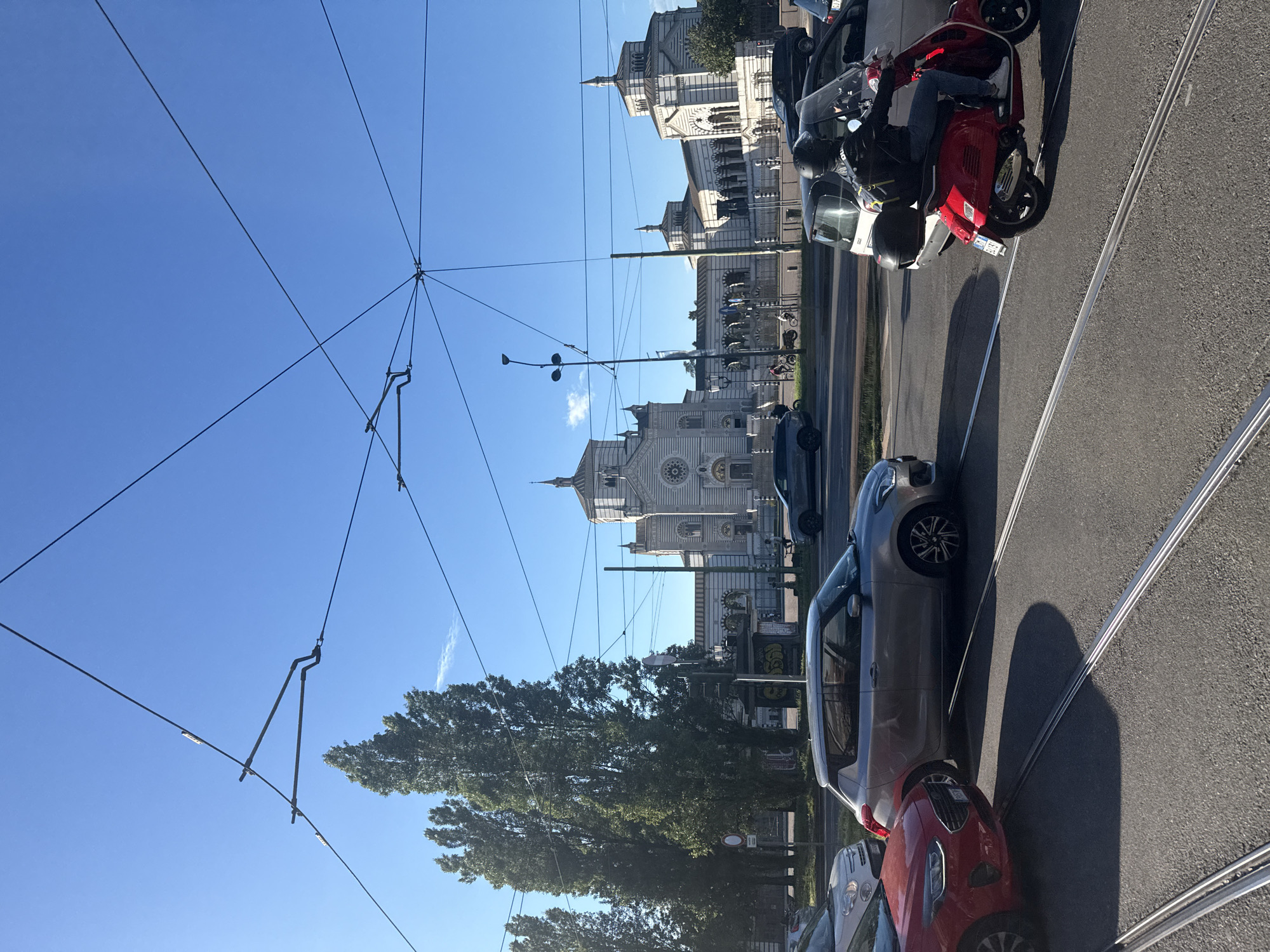 The image depicts a street scene in Milan, Italy. A street lined with parked cars, a red scooter, and tram tracks run along the right side of the frame. Overhead electrical wires intersect against a clear blue sky. In the background, a large, ornate, light-colored building with multiple towers and spires can be seen. Trees line the street, partially obscuring the building. The architecture suggests the Cimitero Monumentale, a major landmark in Milan.