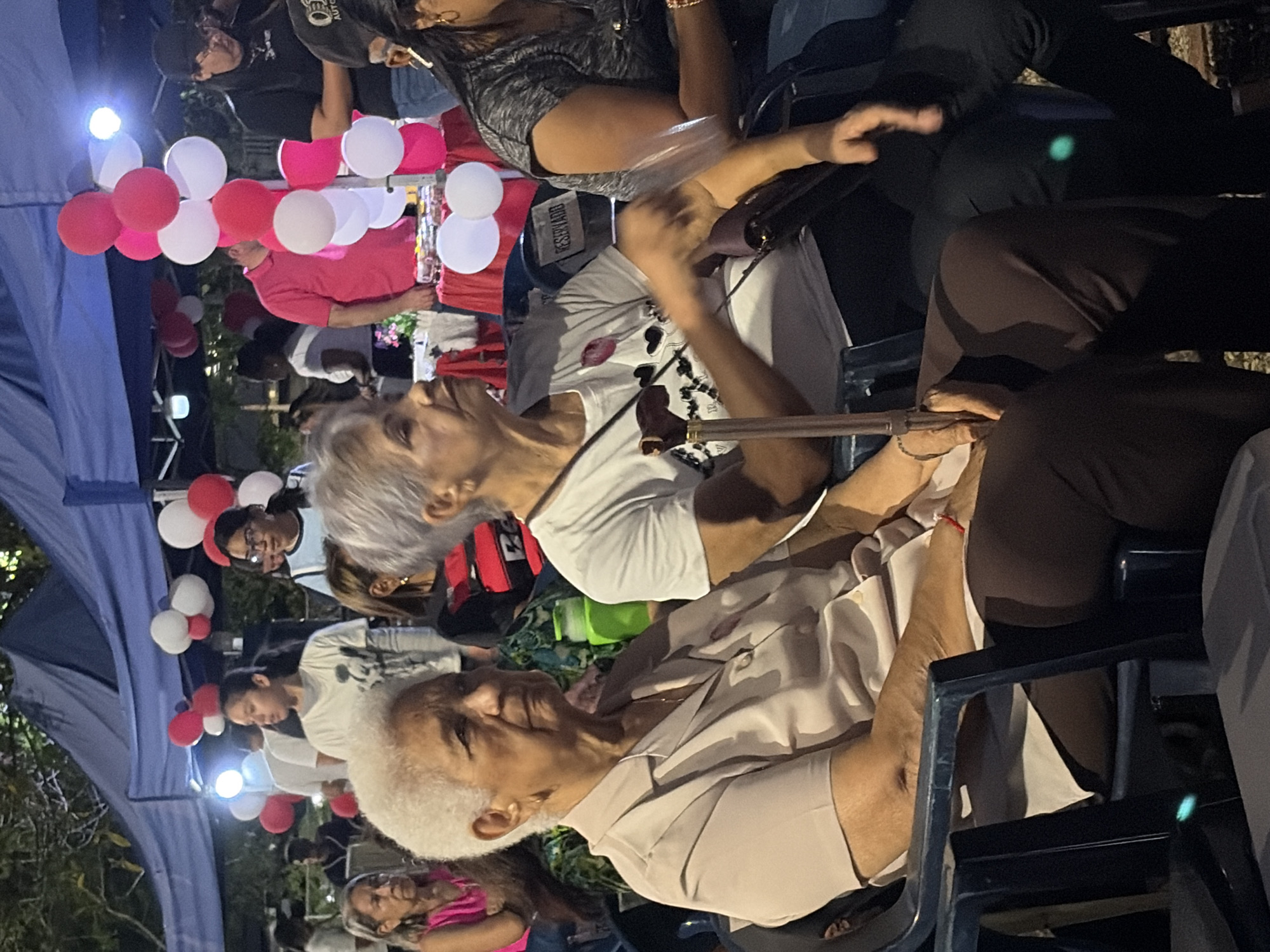 A gathering in Guayana City, Venezuela, includes an elderly woman with white hair wearing a light-colored button-down shirt seated among a crowd. A woman behind her wears a white t-shirt with a black and red design and holds a walking stick. Above them, red and white balloons are tied to a structure. People sit in chairs under a tent.