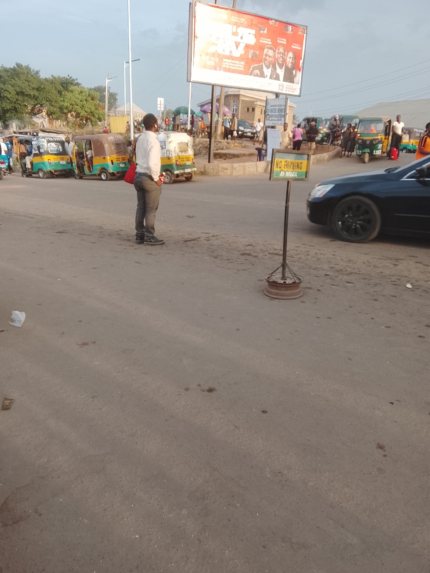 The image depicts a street scene in Gwong, Nigeria. A man in a white shirt and gray pants stands on the side of the road. Several auto rickshaws (tuk-tuks) are parked nearby. A black car is visible to the right, next to a "No Parking" sign. A large billboard is in the background. The road is paved, and there is some litter present. There are other people visible in the background.