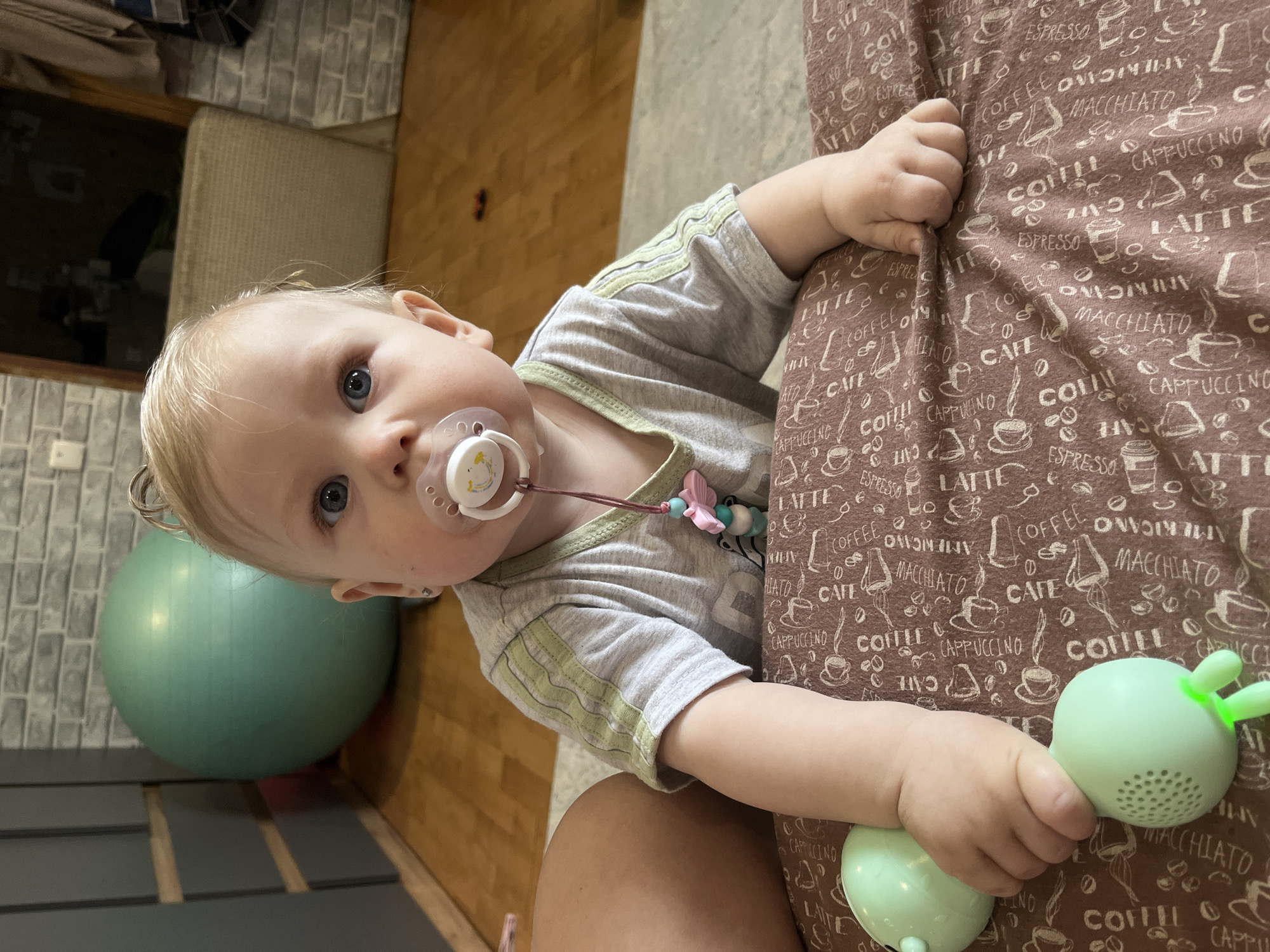 A close-up portrait, taken from a slightly tilted, eye-level perspective, features a fair-skinned baby or toddler with light blonde hair and striking blue eyes. The child wears a light grey t-shirt with horizontal light green stripes on the sleeves and has a white pacifier with yellow details in their mouth, attached to a pacifier clip featuring pink and blue beads and a pink butterfly charm. The baby looks directly at the camera with a curious expression.
One of the baby's hands is clutching a textured brown fabric, which is extensively patterned with coffee-related text and illustrations such as "LATTE", "ESPRESSO", "COFFEE", "MACCHIATO", "CAPPUCCINO", "AMERICANO", and "CAFE". The other hand holds a light green toy, shaped like an elongated sphere with speaker-like perforations and two prominent, ear-like protrusions at one end. Part of an adult's bare leg is visible at the bottom right, suggesting the baby is sitting on or next to them.
The background reveals an indoor setting with a large, teal exercise ball positioned on a light wooden floor. Beyond the wooden floor, a section of grey-toned, possibly tiled or brick-patterned, flooring is visible. The scene captures a peaceful, domestic moment in Vesyoloye, Russia.