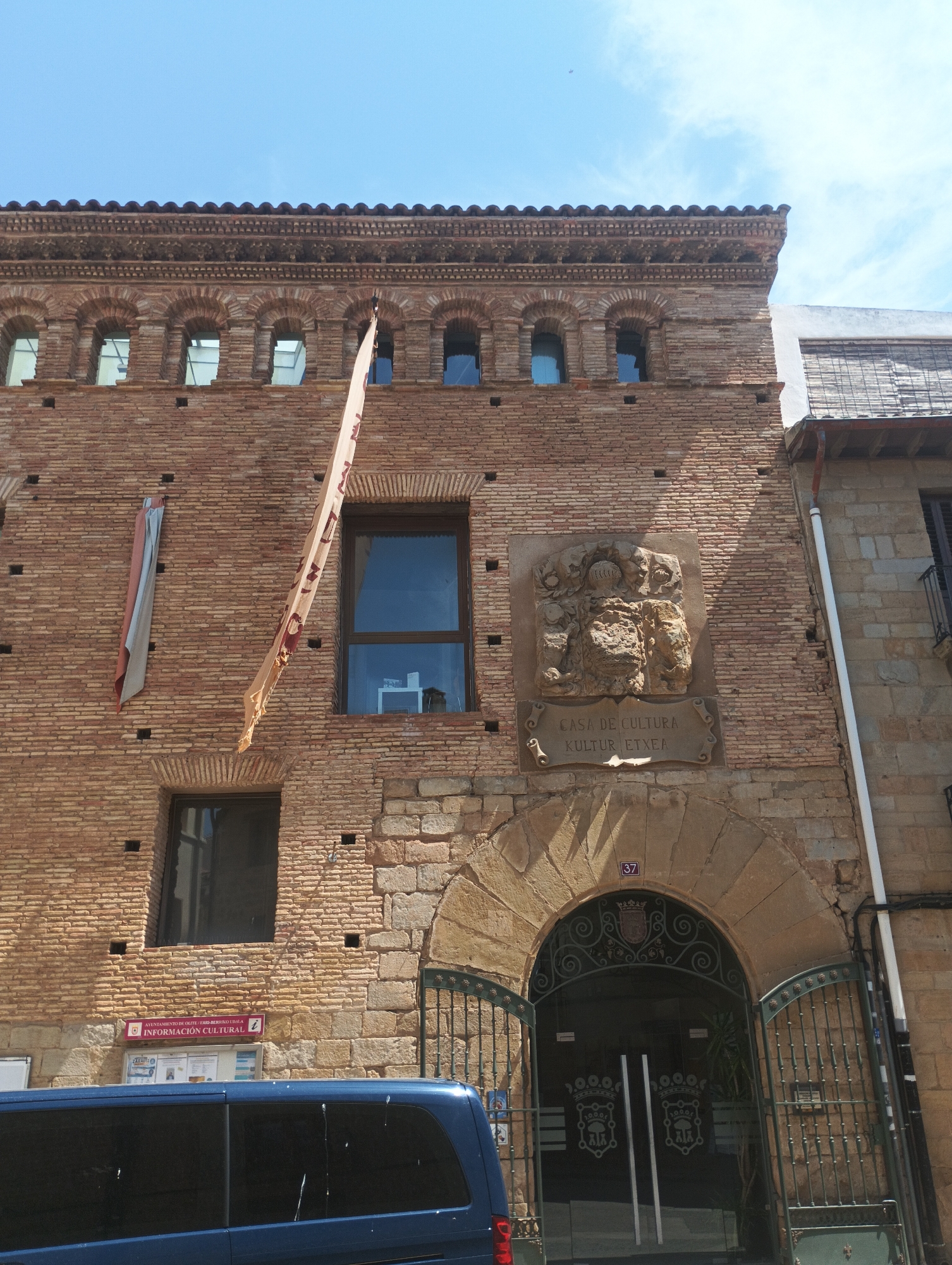A medium shot captures the facade of an old brick building in a city. The building features an arched entryway with an open metal gate revealing a glimpse of the interior. Above the entrance, a stone carving depicts a coat of arms, with the inscription "CASA DE CULTURA / KULTUR ETXEA" below it. Several windows are visible on the upper floors, and a banner hangs from the facade, partially obscuring one of the windows. A blue van is parked in front of the building. The sky above is clear blue with scattered clouds. A small number "37" is visible above the arched entrance. The building is made of weathered red brick with decorative brickwork along the roofline and around some of the windows. A smaller, faded banner hangs on the left side of the building.