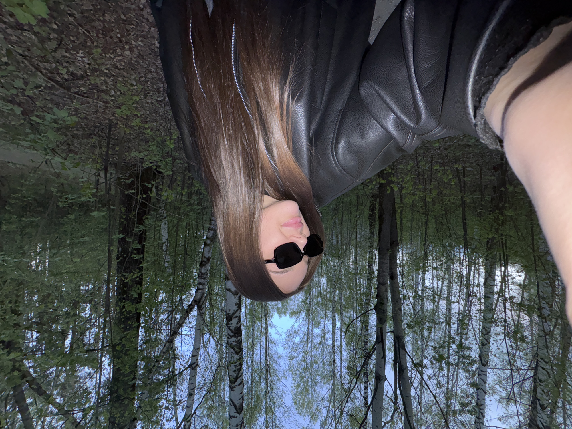 A woman is wearing a black leather jacket and black sunglasses in a forest or wooded area near Chaikovsky, Russia. The woman is facing downwards. The background shows a dense forest with birch trees. The lighting suggests it is either dusk or dawn.
