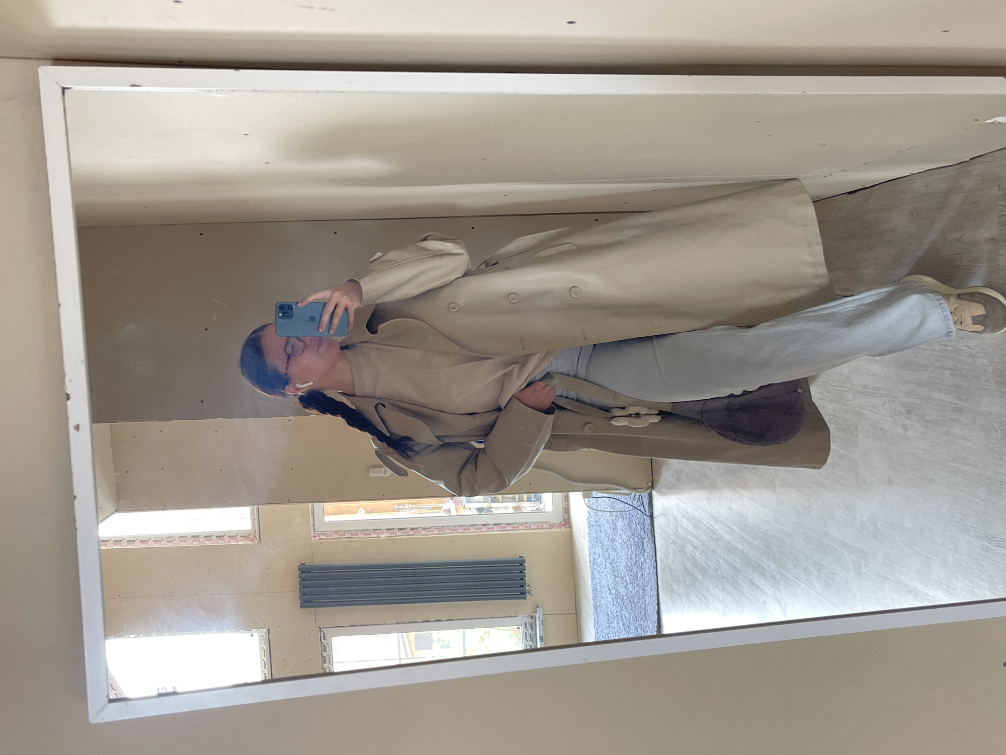 A person is taking a selfie in front of a rectangular mirror. The person is wearing a beige trench coat, light blue jeans, and beige shoes. They are holding a blue smartphone in their left hand, and their face is visible in the mirror. The background shows a room undergoing renovations, with unfinished drywall, windows, and a radiator. The location is likely Мосметаллоконструкция Онт, Russia.