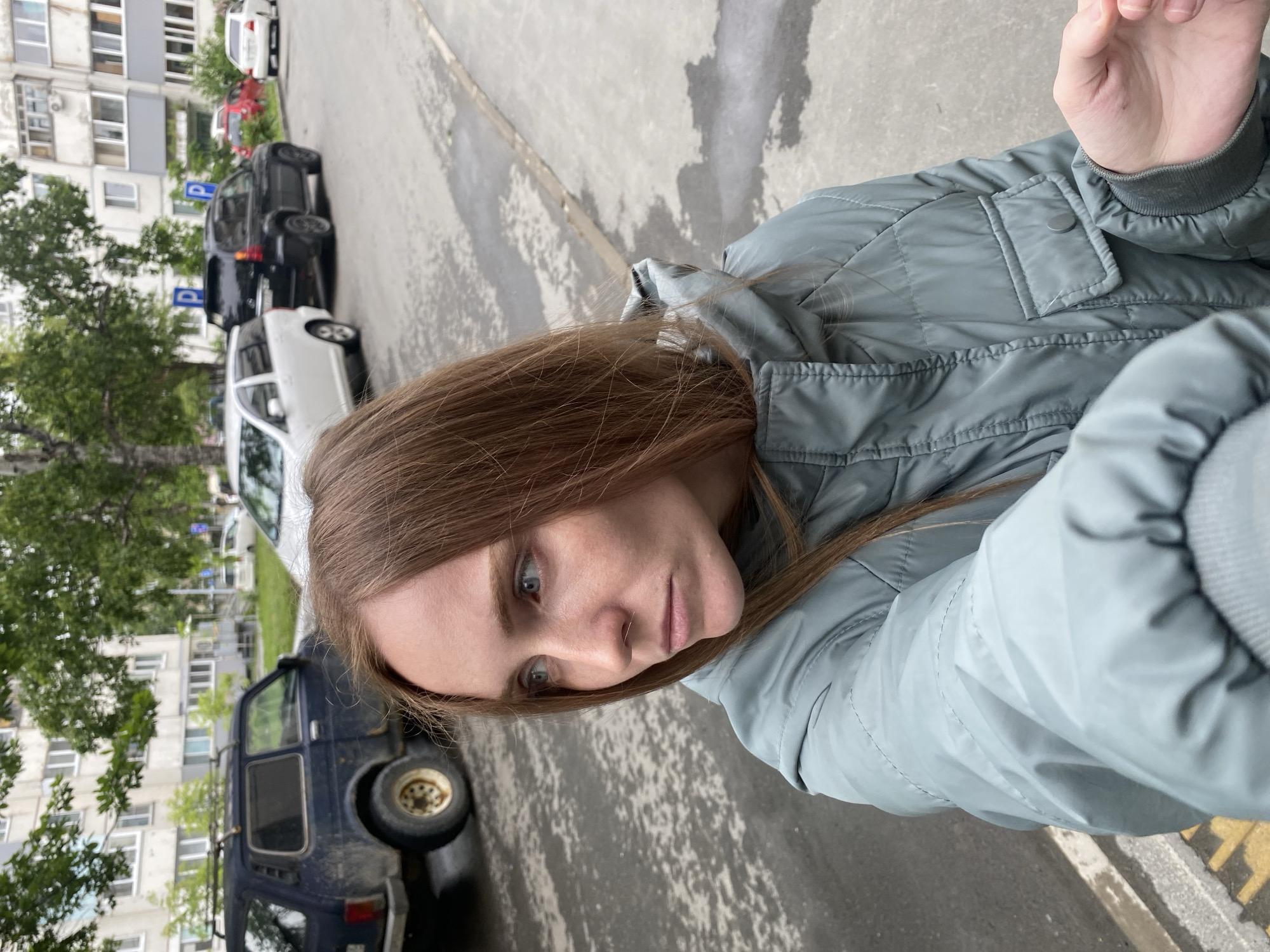 The image shows a young woman with long brown hair and blue eyes standing on a street in Yuzhno-Sakhalinsk, Russia. She is wearing a light blue puffy jacket and looking to her left. In the background, there are several parked cars, including a black SUV, a white hatchback, and a dark blue older model SUV. A multi-story apartment building is visible behind the cars, with trees and foliage in front of it. The street appears to be wet, suggesting it may have recently rained. The scene is captured from a close-up perspective, with the woman's face and upper body occupying most of the frame.